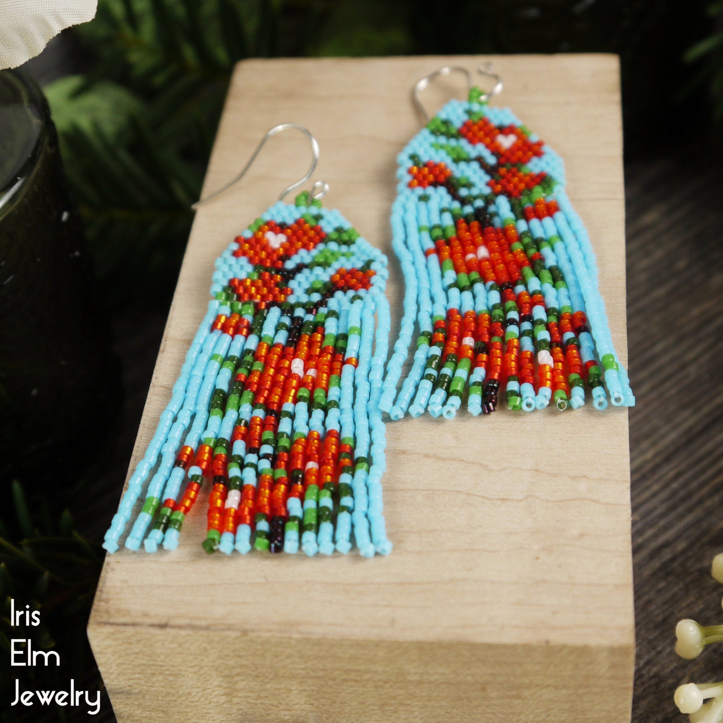 Colorful beaded light blue and red flower-patterned seed bead fringe earrings on a wooden block with 'Iris Elm Jewelry' branding.