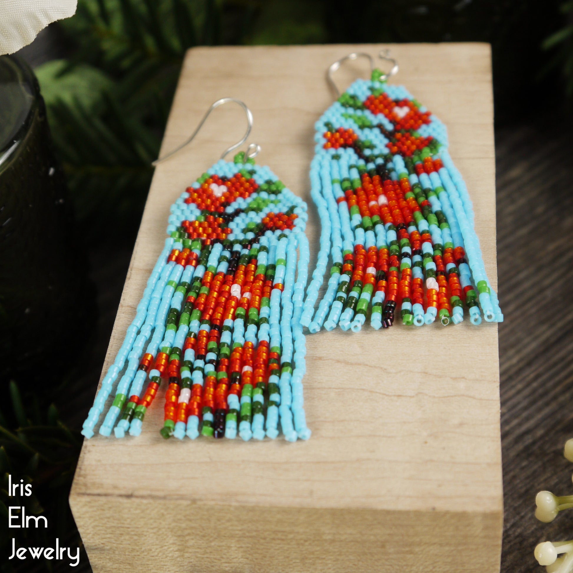 Colorful beaded light blue and red flower-patterned seed bead fringe earrings on a wooden block with 'Iris Elm Jewelry' branding.