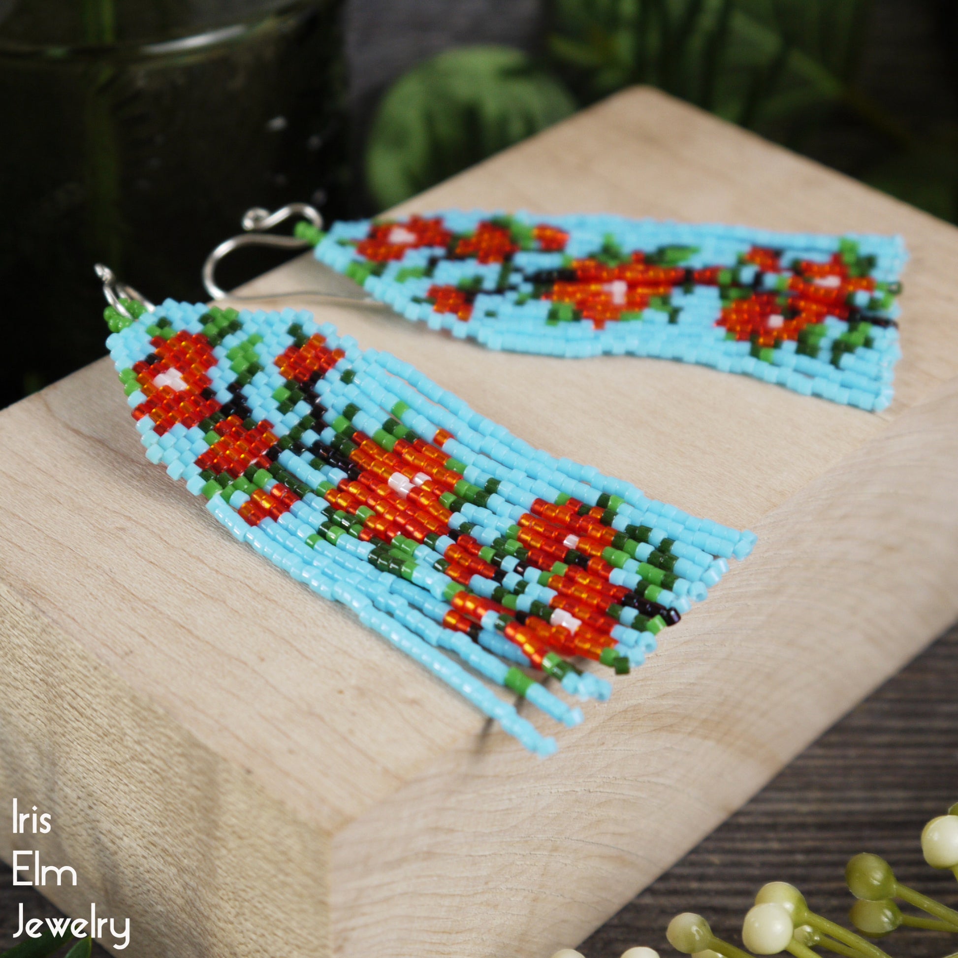 Beaded light blue handmade fringe earrings with red floral design on a wooden surface, brand 'Iris Elm Jewelry' visible.