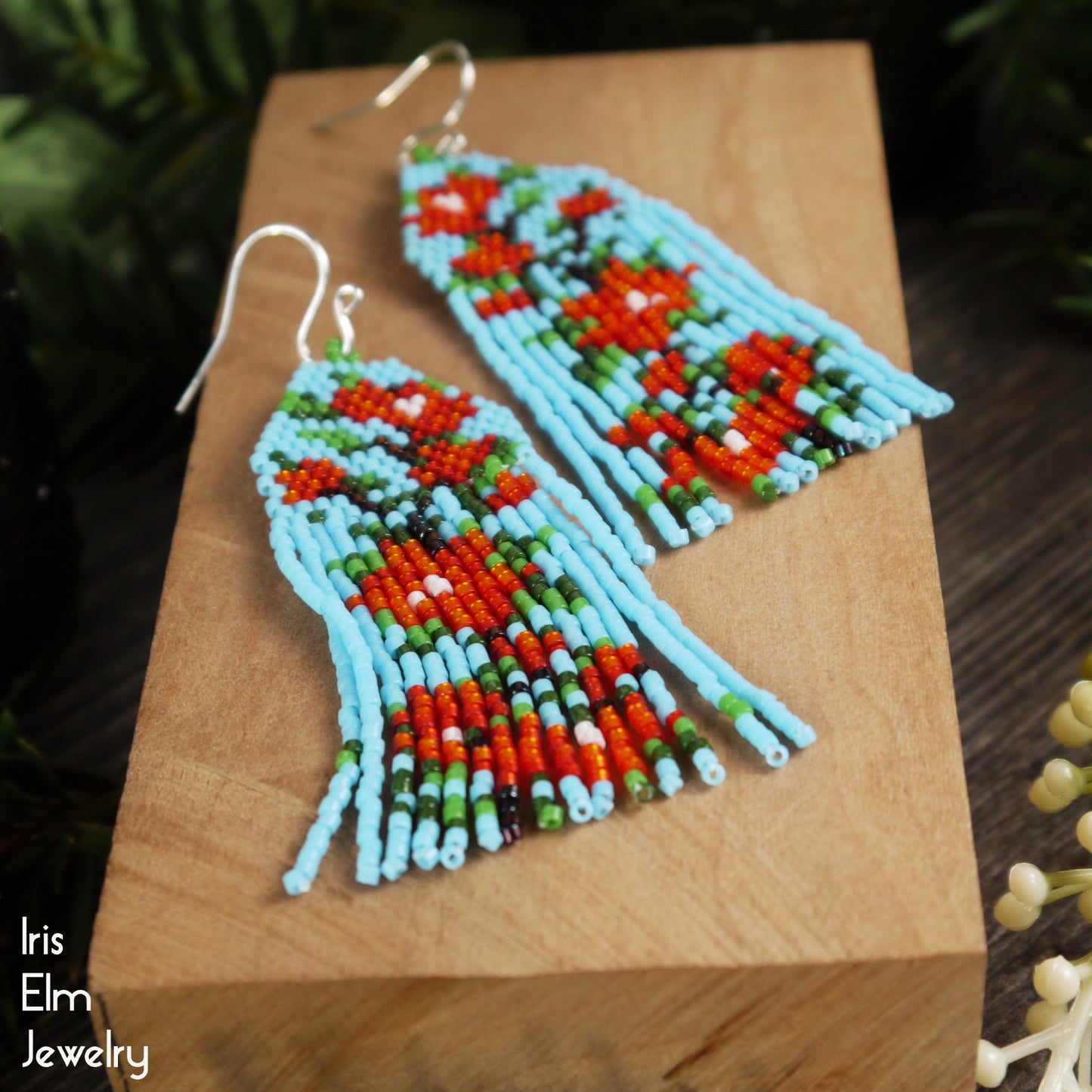 Colorful beaded red flower seed bead fringe earrings on a wooden block with 'Iris Elm Jewelry' branding.