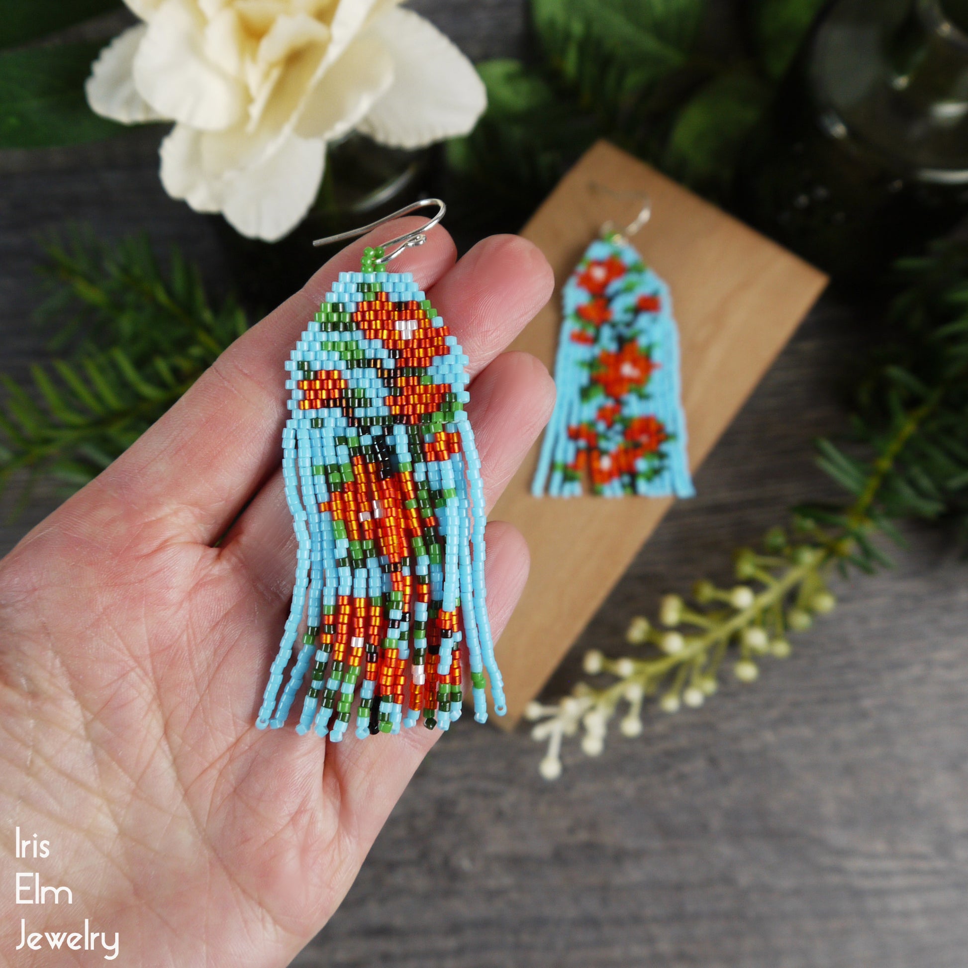 Beaded light blue and red orange floral fringe earrings held in a hand with a floral background, featuring the brand 'Iris Elm Jewelry'.