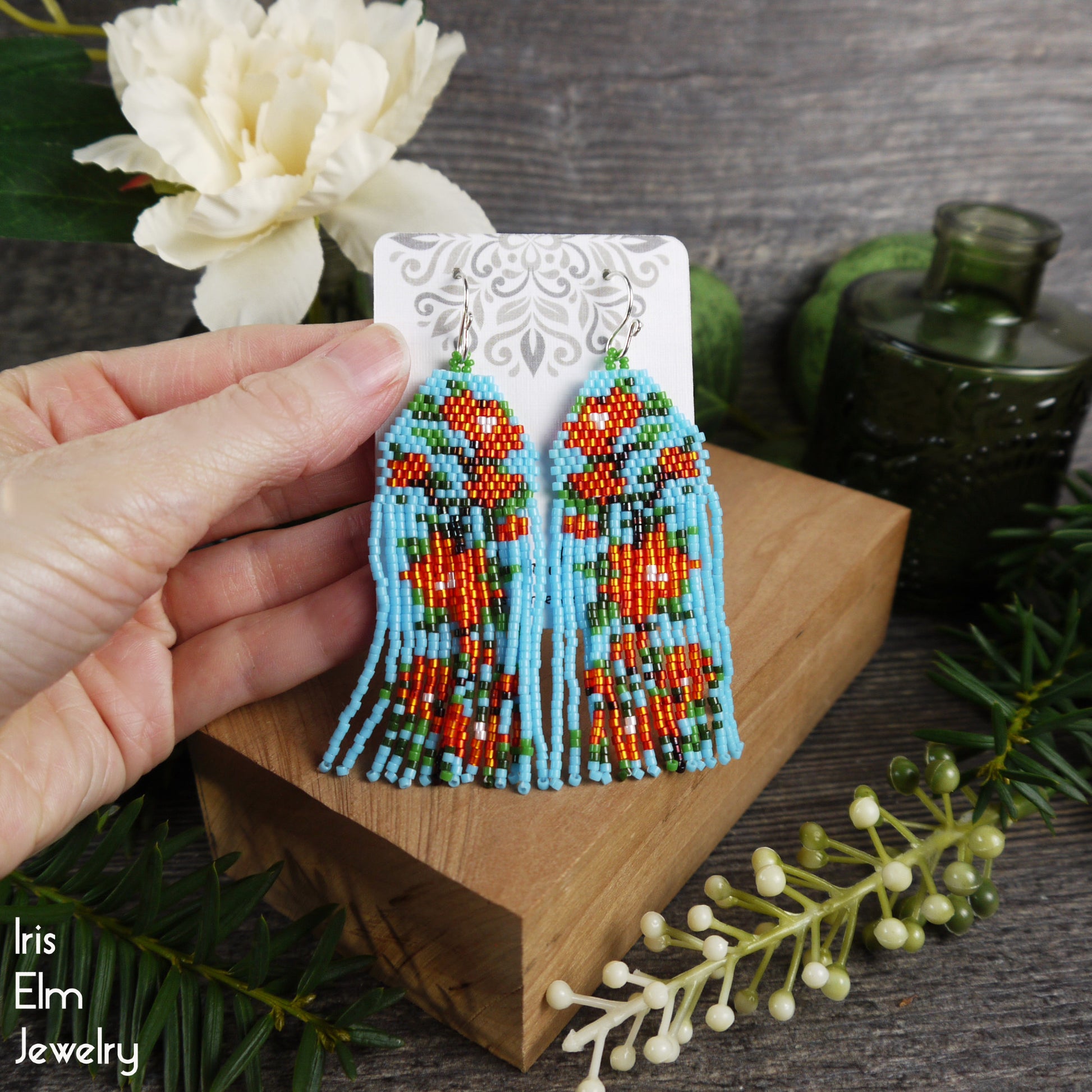 Hand holding a pair of beaded light blue and red fringe earrings on a wooden block with a decorative background