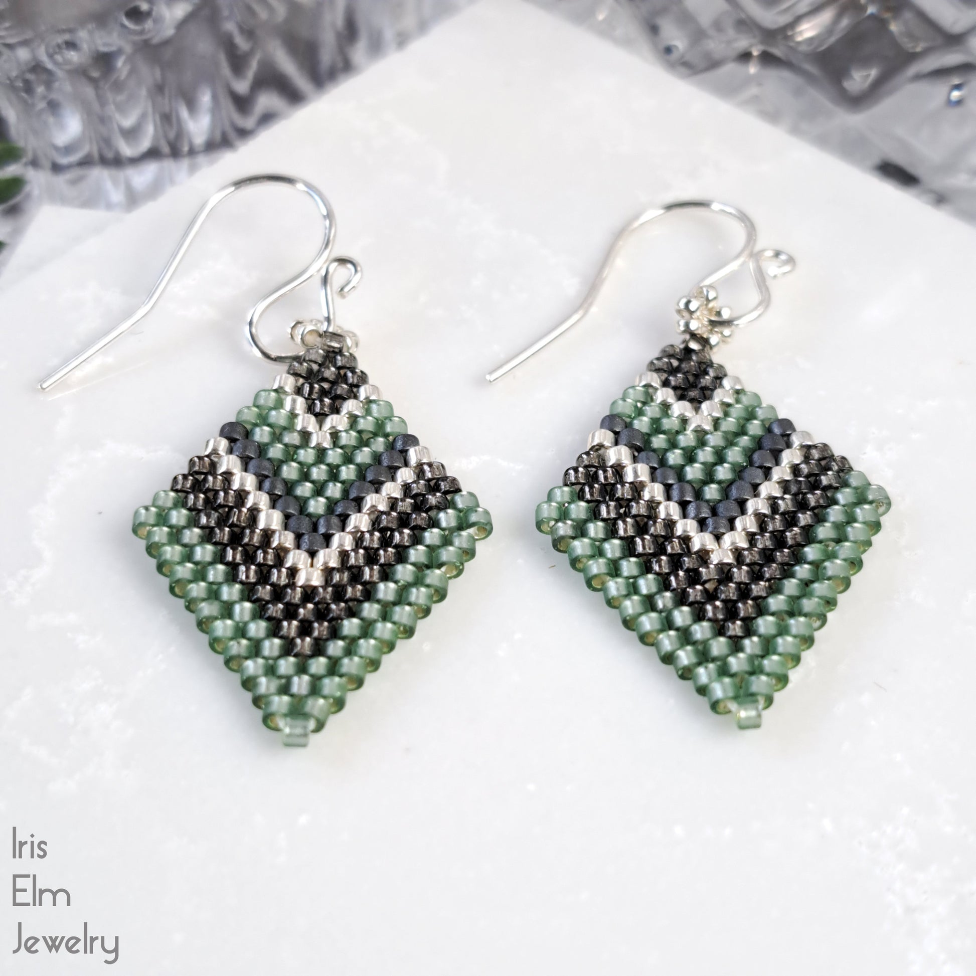 Pair of beaded earrings with green and grey chevron diamond pattern on a white surface, branded 'Iris Elm Jewelry'.