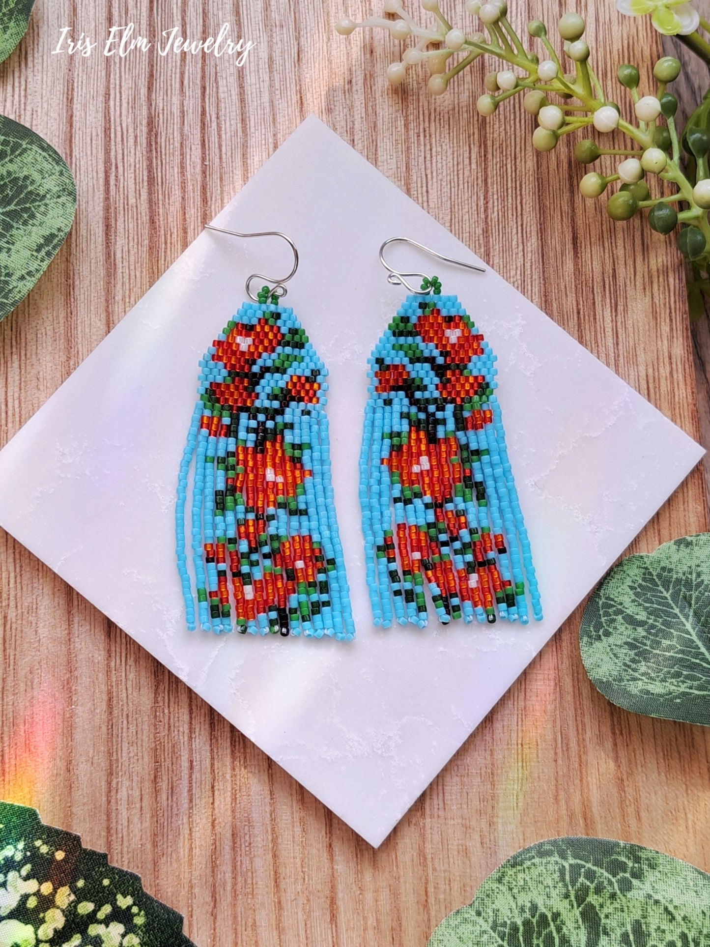 Beaded red cascading flower light blue seed bead fringe earrings on a white square with green leaves around