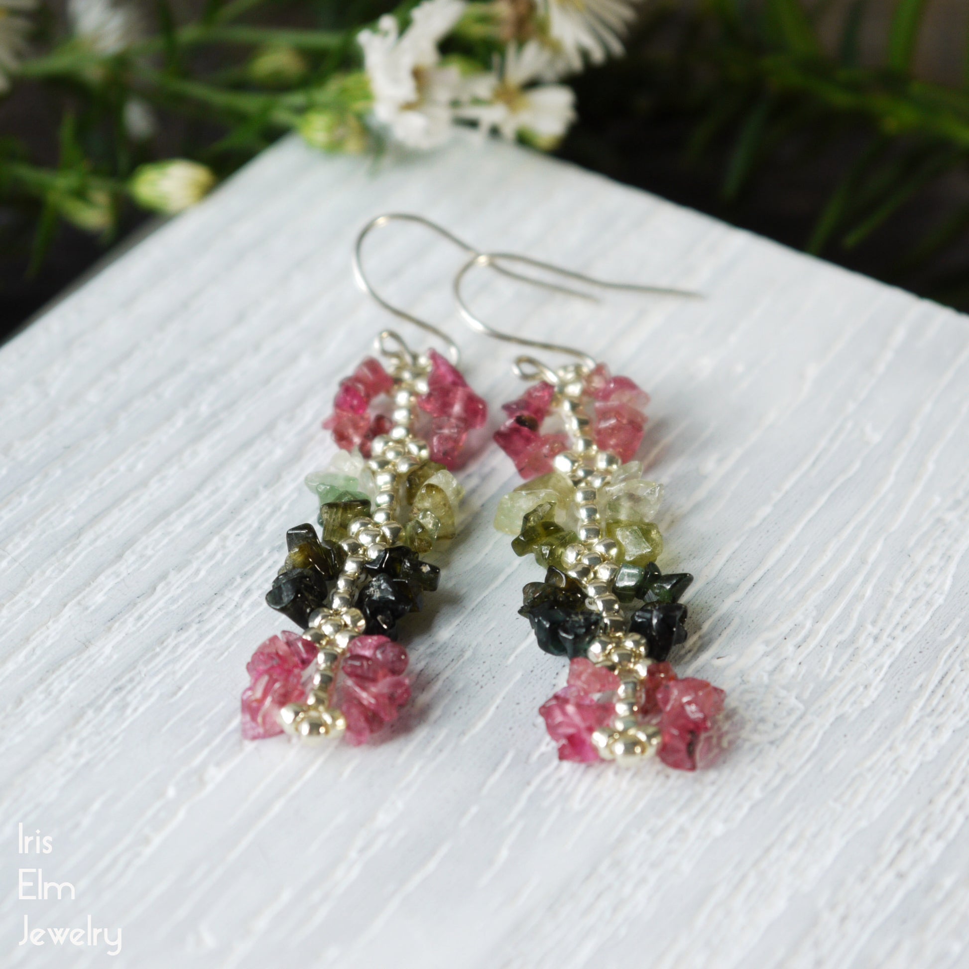 Jewelry earrings with multicolored stone chip beads on a white surface with flowers in the background