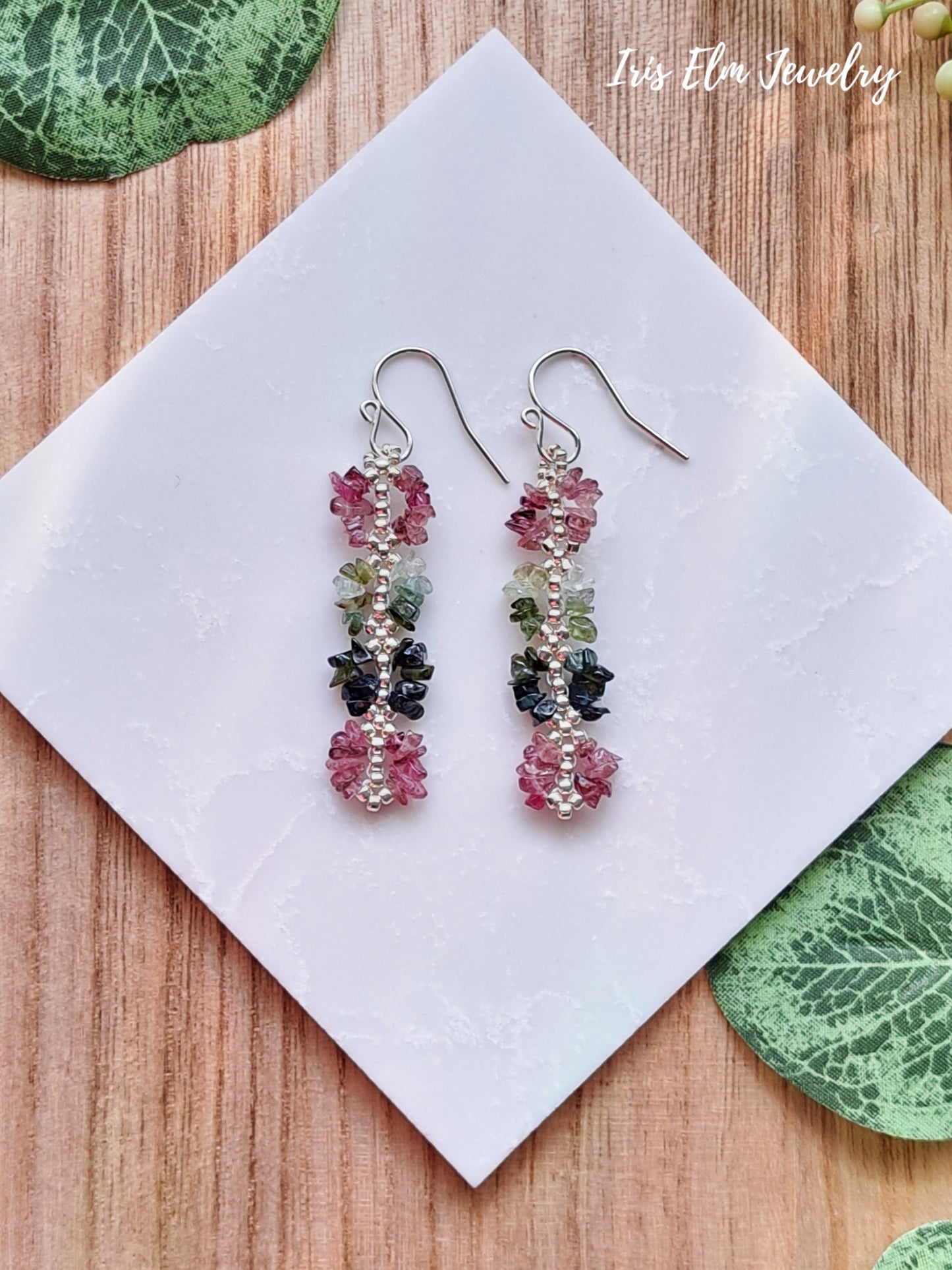 Pair of colorful beaded pink and green rainbow tourmaline gemstone chip earrings on a white card with 'Iris Elm Jewelry' branding, placed on a wooden surface.