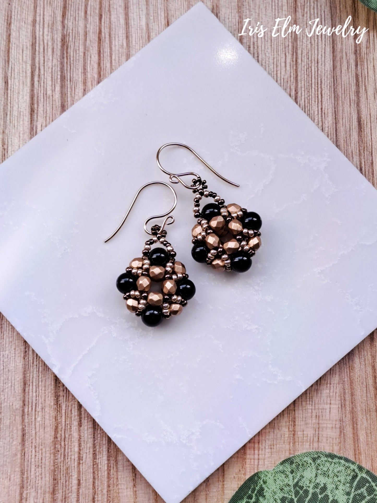 Pair of black onyx and gold glass beaded earrings on a white tile with 'Iris Elm Jewelry' branding.