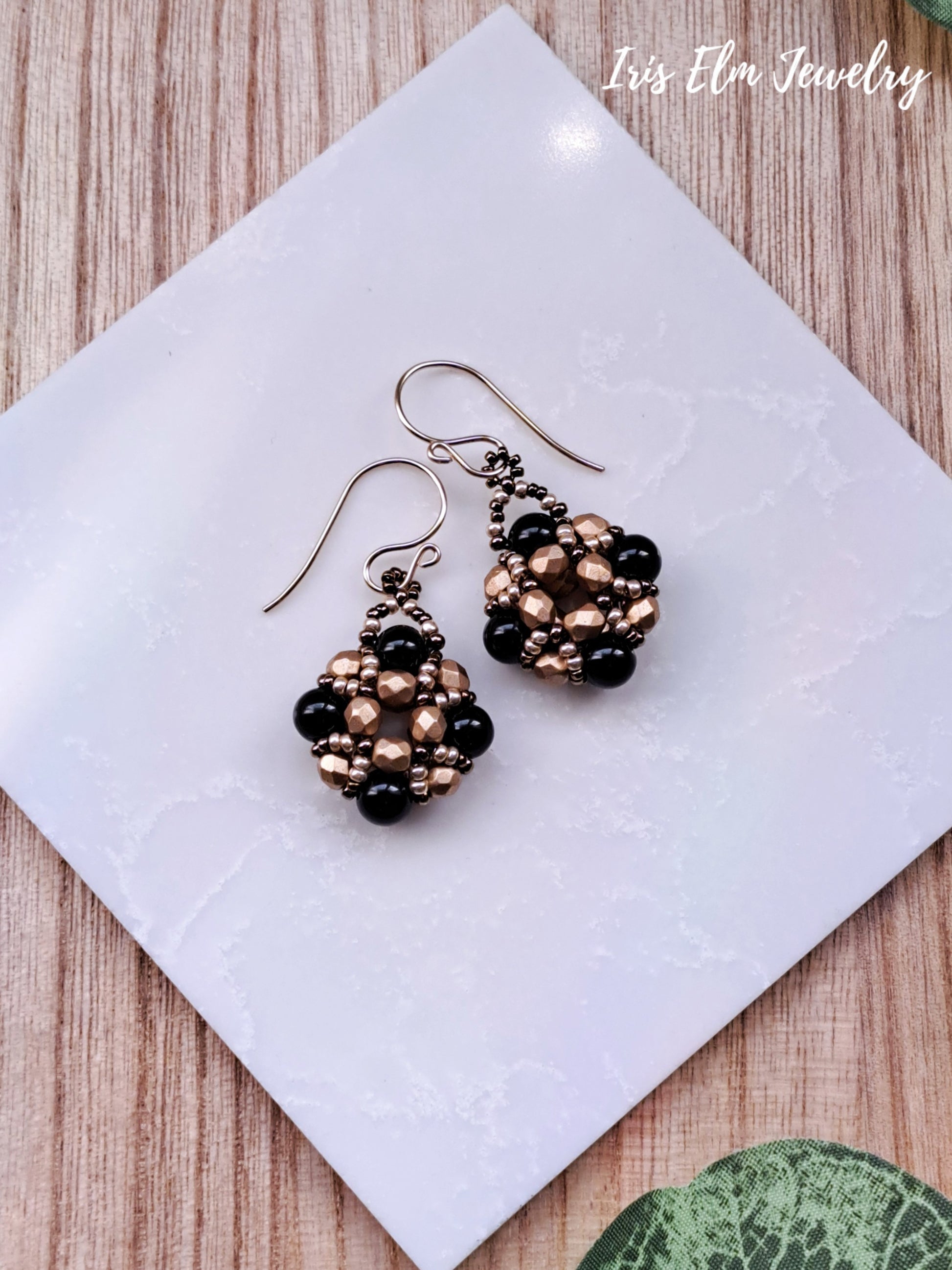Pair of black onyx and gold glass beaded earrings on a white tile with 'Iris Elm Jewelry' branding.