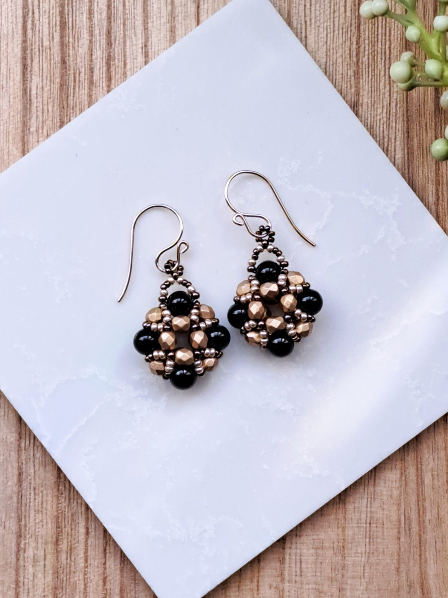 Pair of beaded handmade black onyx gemstone and gold glass earrings on a white tile with 'Iris Elm Jewelry' branding.