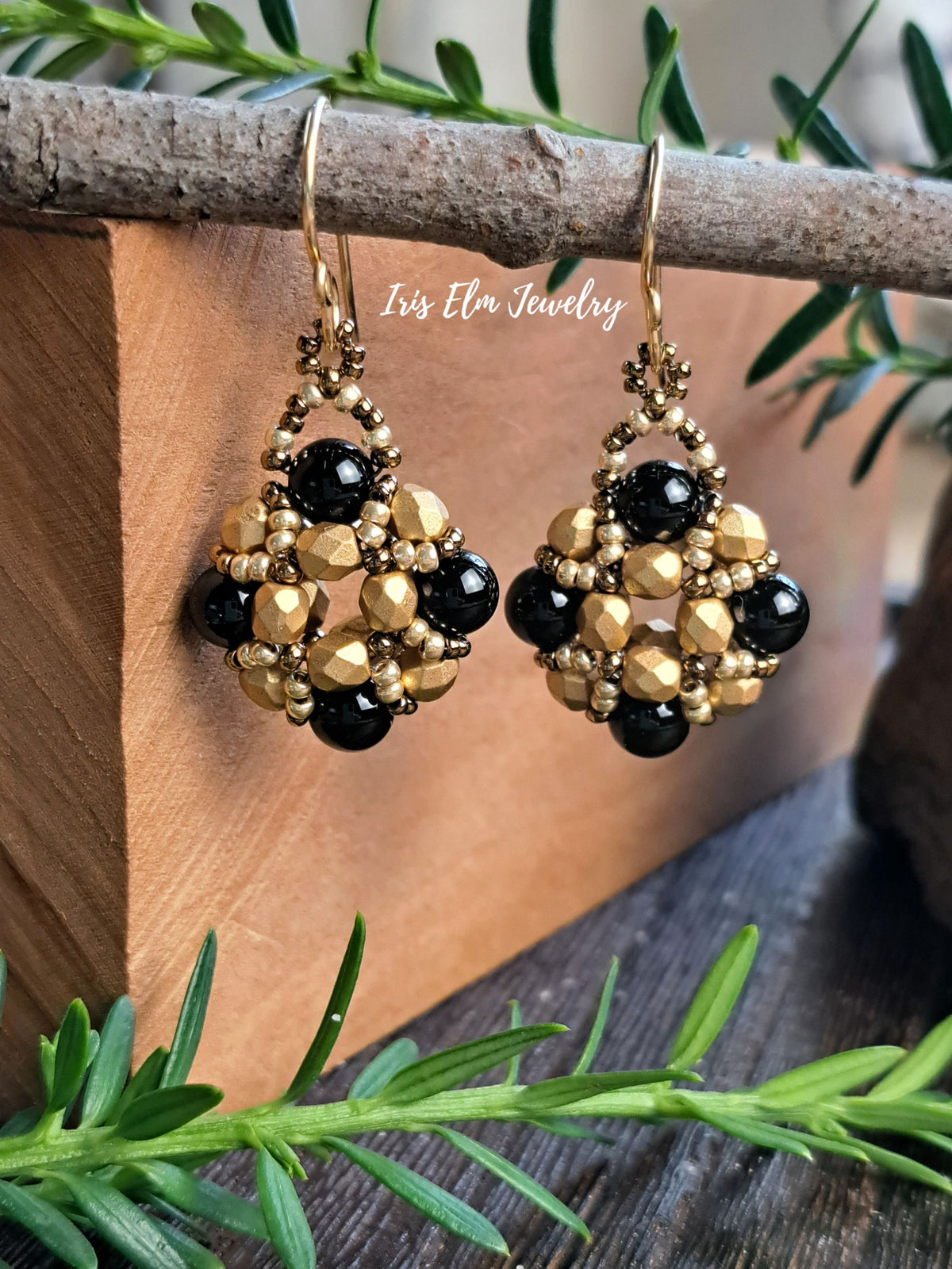 A pair of handmade beaded dangle earrings with black onyx and gold beads, hanging from a rustic wooden branch. The background features a wooden block and green pine branches, with a watermark reading 'Iris Elm Jewelry' across the center.