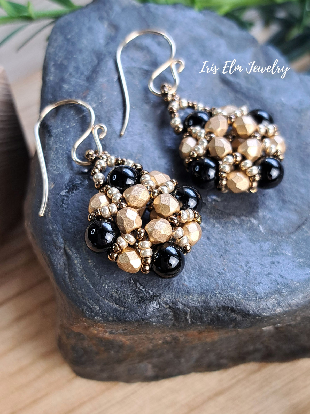 A pair of beaded dangle earrings with black onyx and metallic gold faceted beads and silver-toned fishhook wires, displayed on a dark stone surface. The text 'Iris Elm Jewelry' is visible in the upper right.