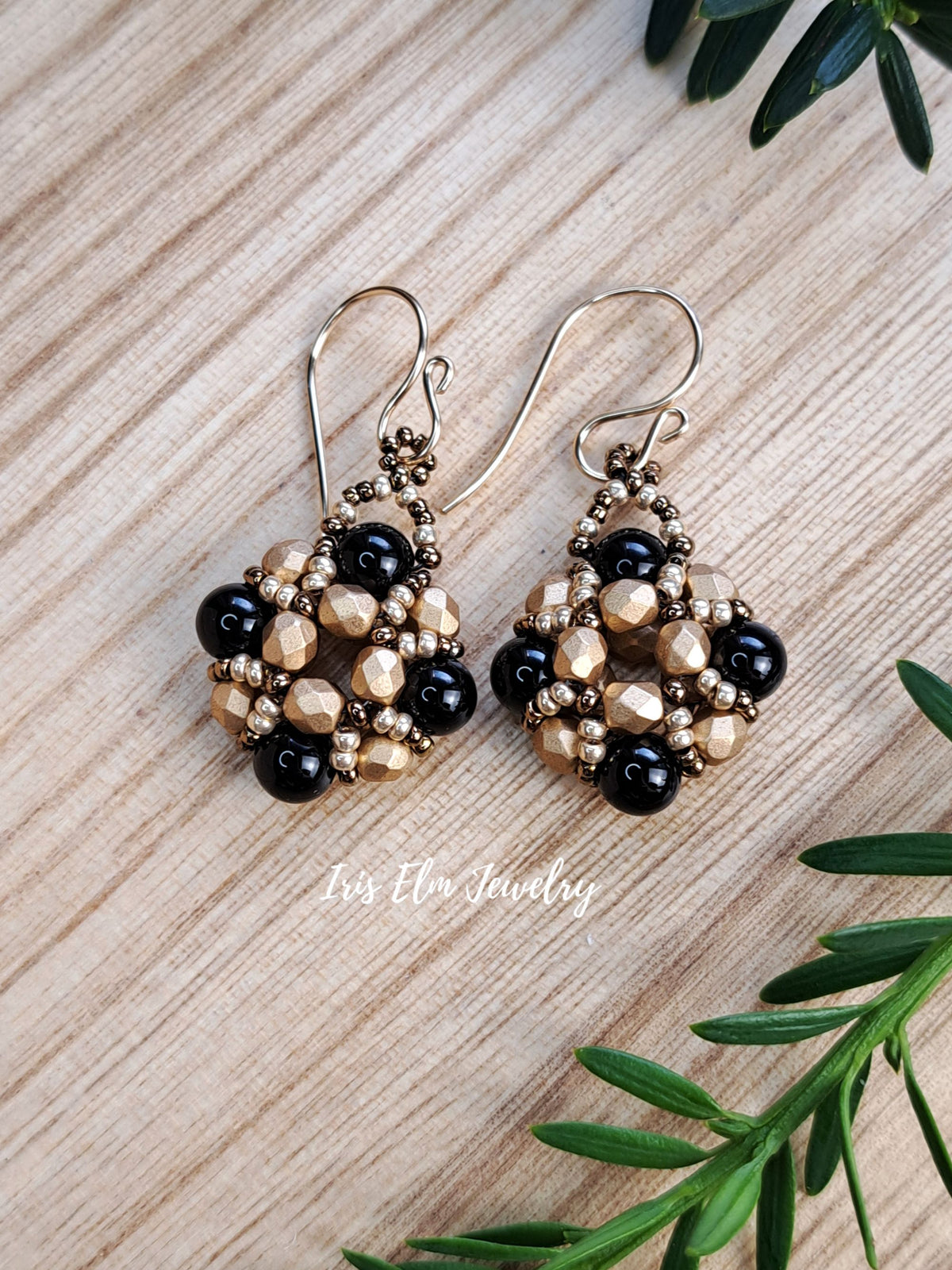 A pair of beaded dangle earrings with black onyx and metallic gold beads, displayed on a light wood background with green leaves in the corners. The brand name 'Iris Elm Jewelry' is watermarked near the bottom.