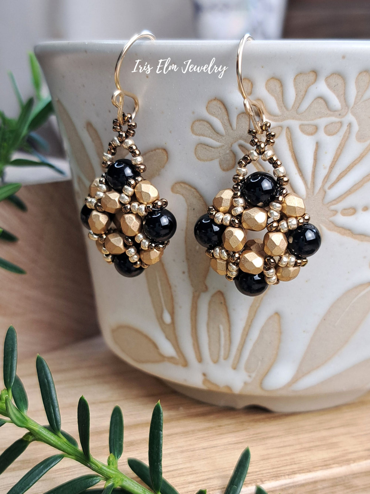 A pair of gold and black beaded dangle earrings displayed on a ceramic cup, with green pine branches in the foreground and a wooden surface underneath. The brand name 'Iris Elm Jewelry' is watermarked near the top.