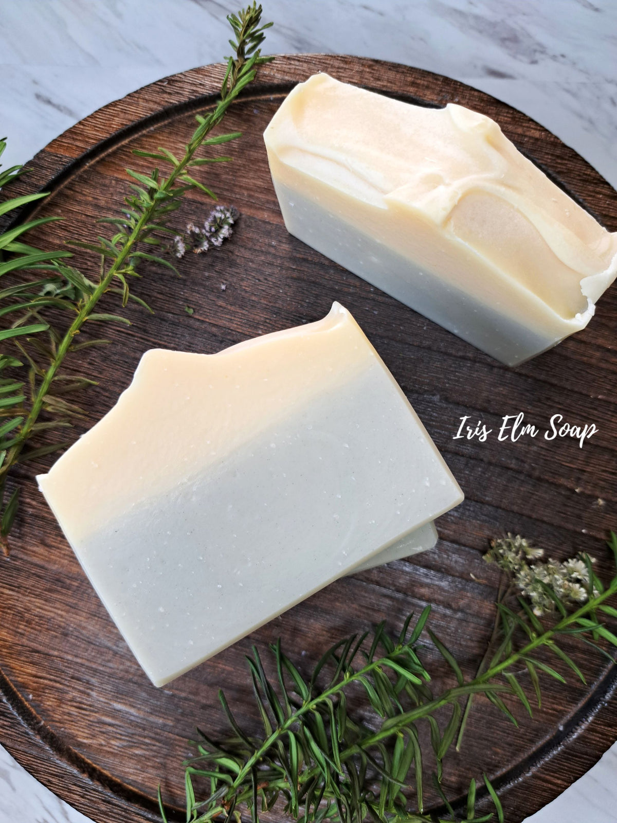 Two rectangular bars of cedarwood lavender essential oil handmade soap with a white-to-cream gradient, styled on a dark wooden board with green rosemary sprigs and a white marble surface. The brand name 'Iris Elm Soap' is printed on the wood.