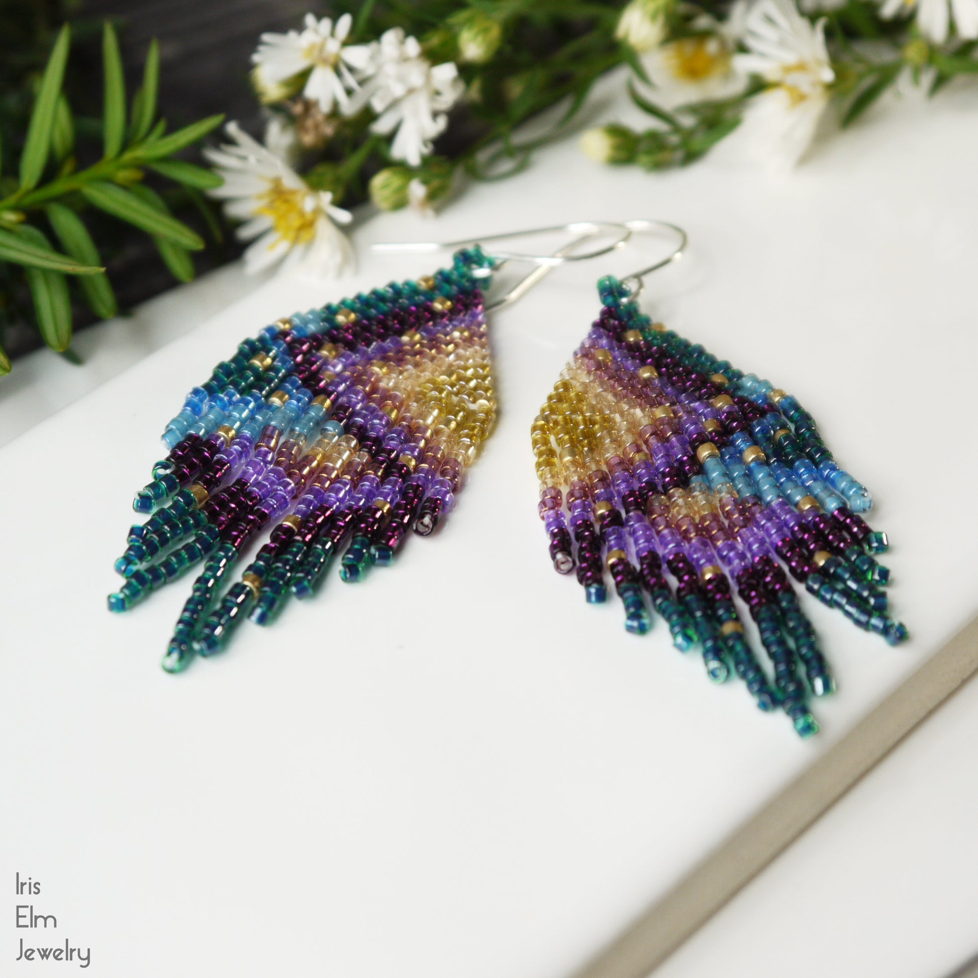 Colorful beaded green, purple and blue seed bead fringe earrings on a white surface with flowers in the background