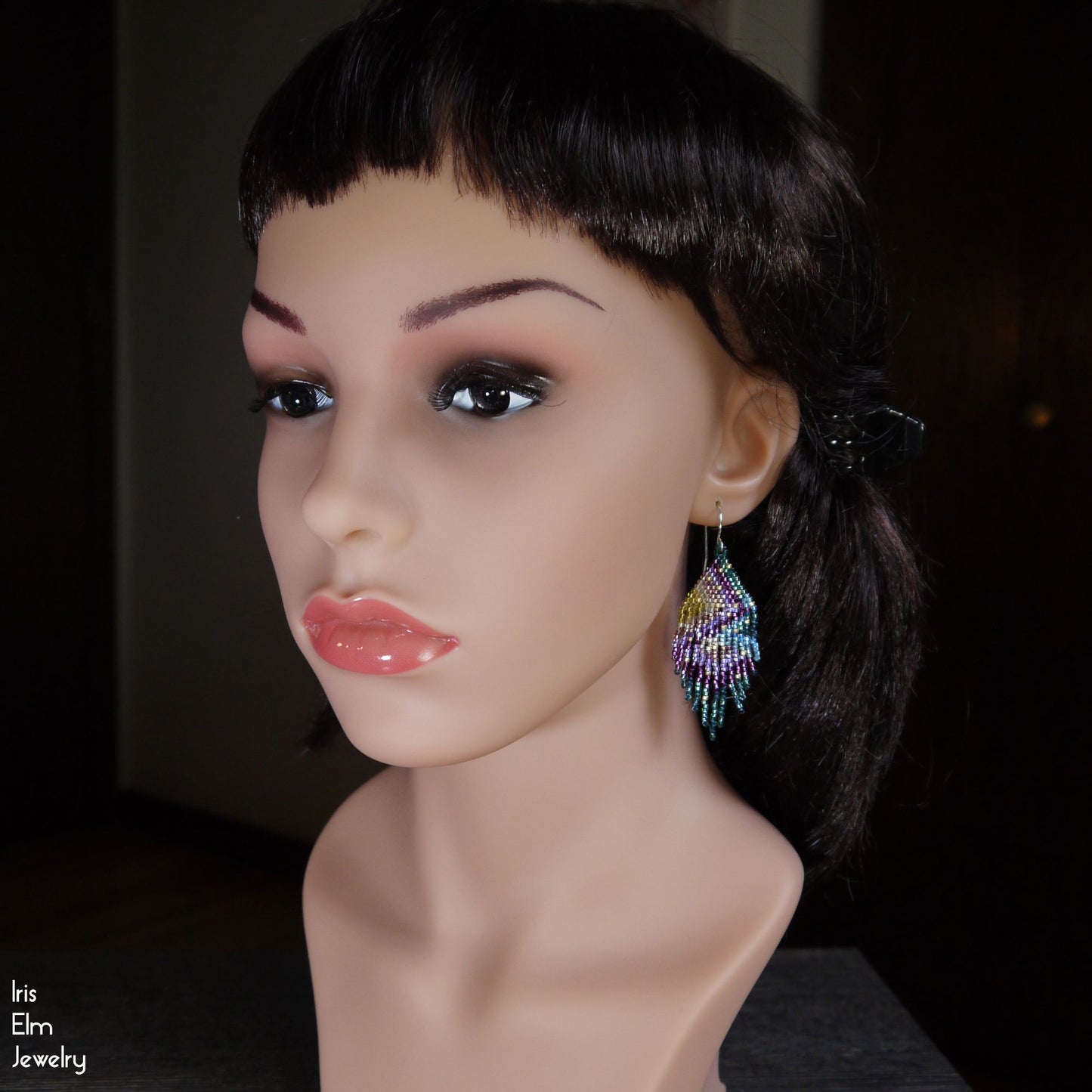 Mannequin head with colorful beaded purple and green rainbow fringe earrings on a dark background