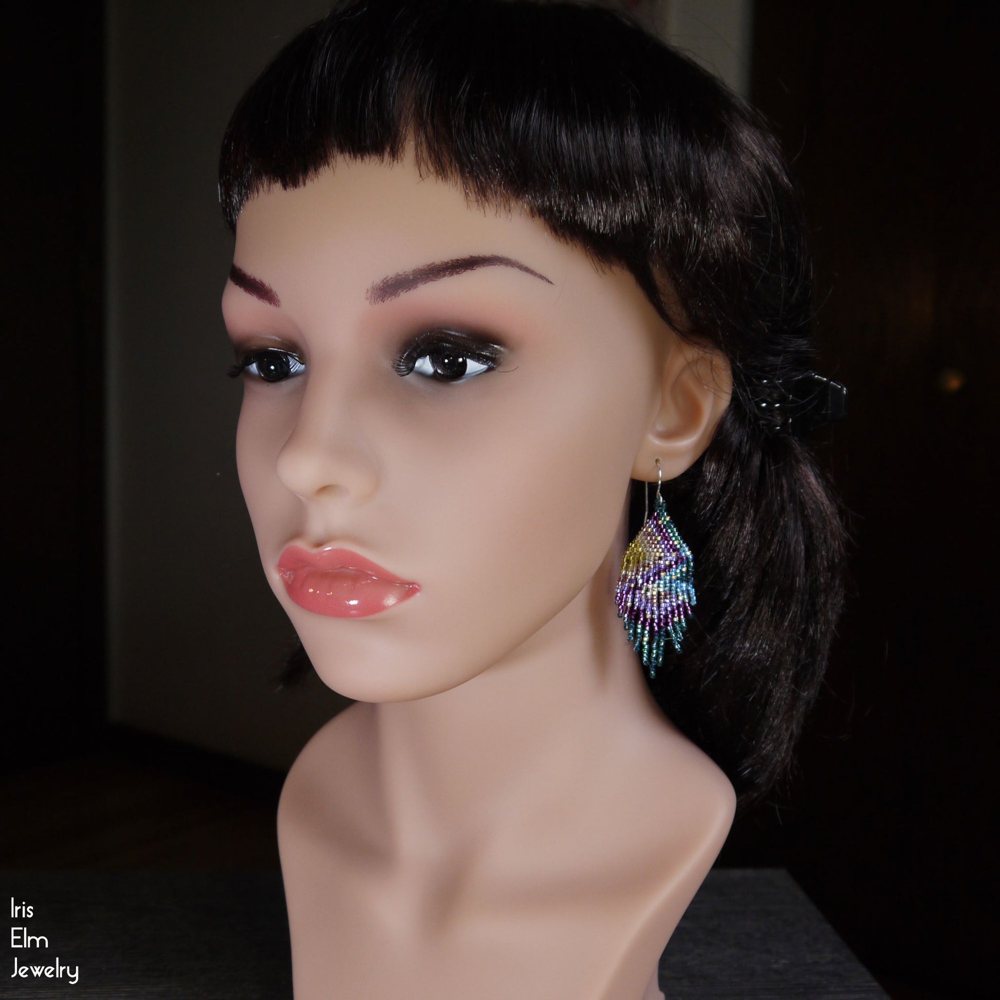 Mannequin head with colorful beaded purple and green rainbow fringe earrings on a dark background