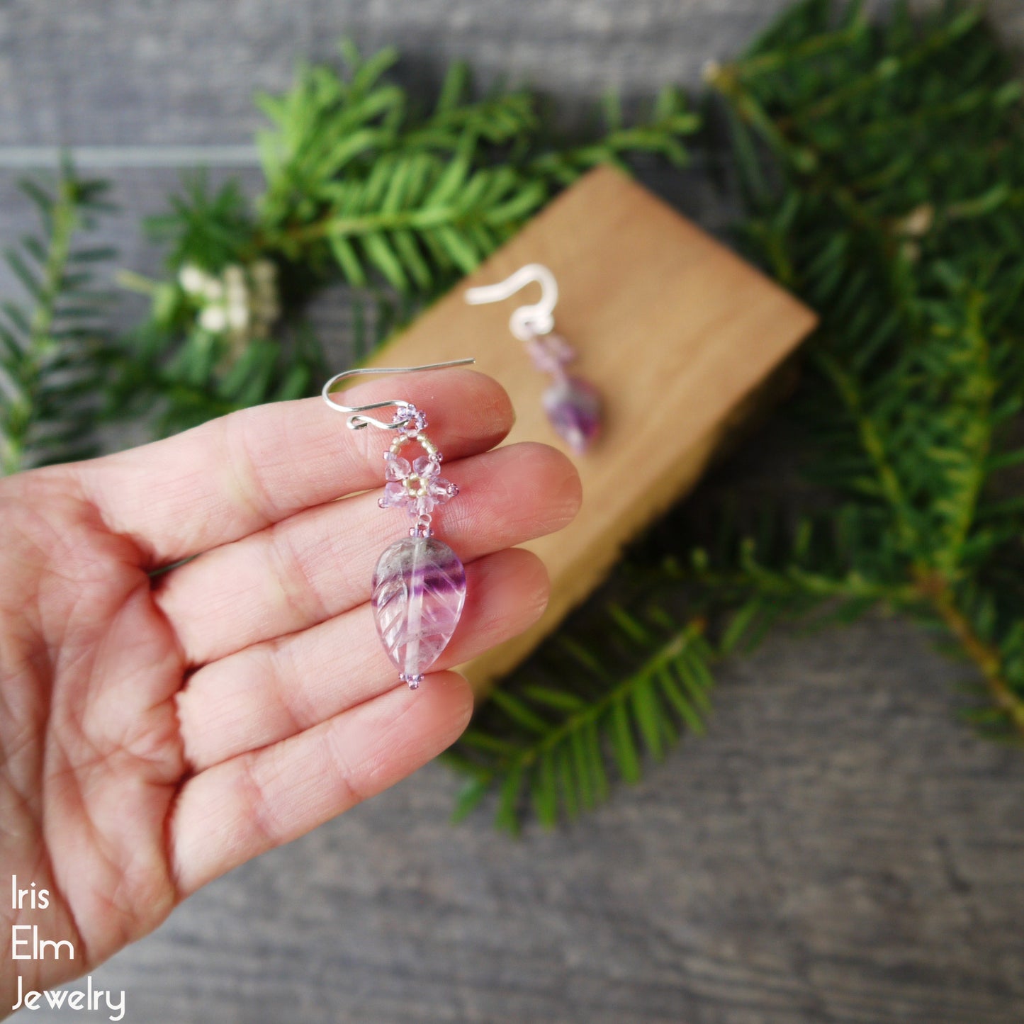 Hand holding a pair of purple leaf-shaped fluorite and amethyst beaded flower earrings with a blurred greenery background