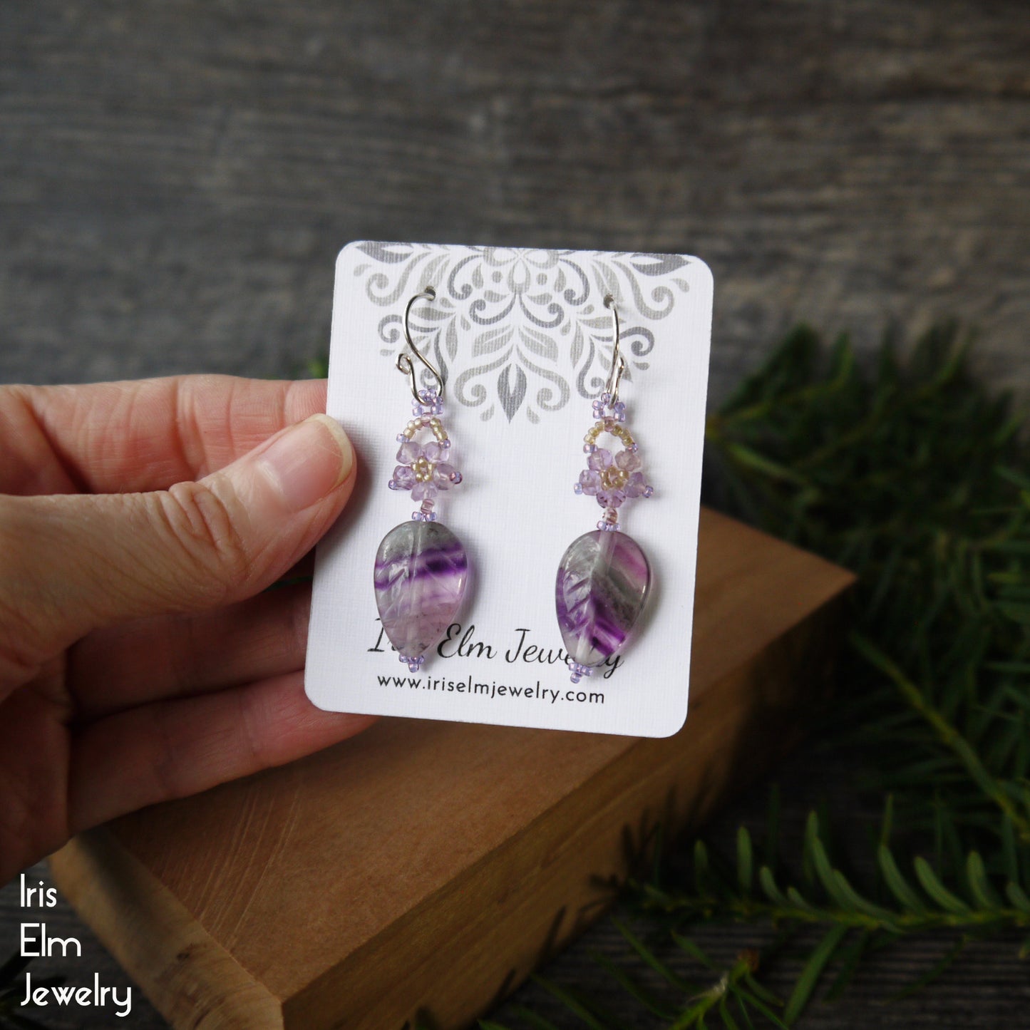 rainbow fluorite and amethyst floral leaf Earrings on a 'Iris Elm Jewelry' card held by a hand with a natural background