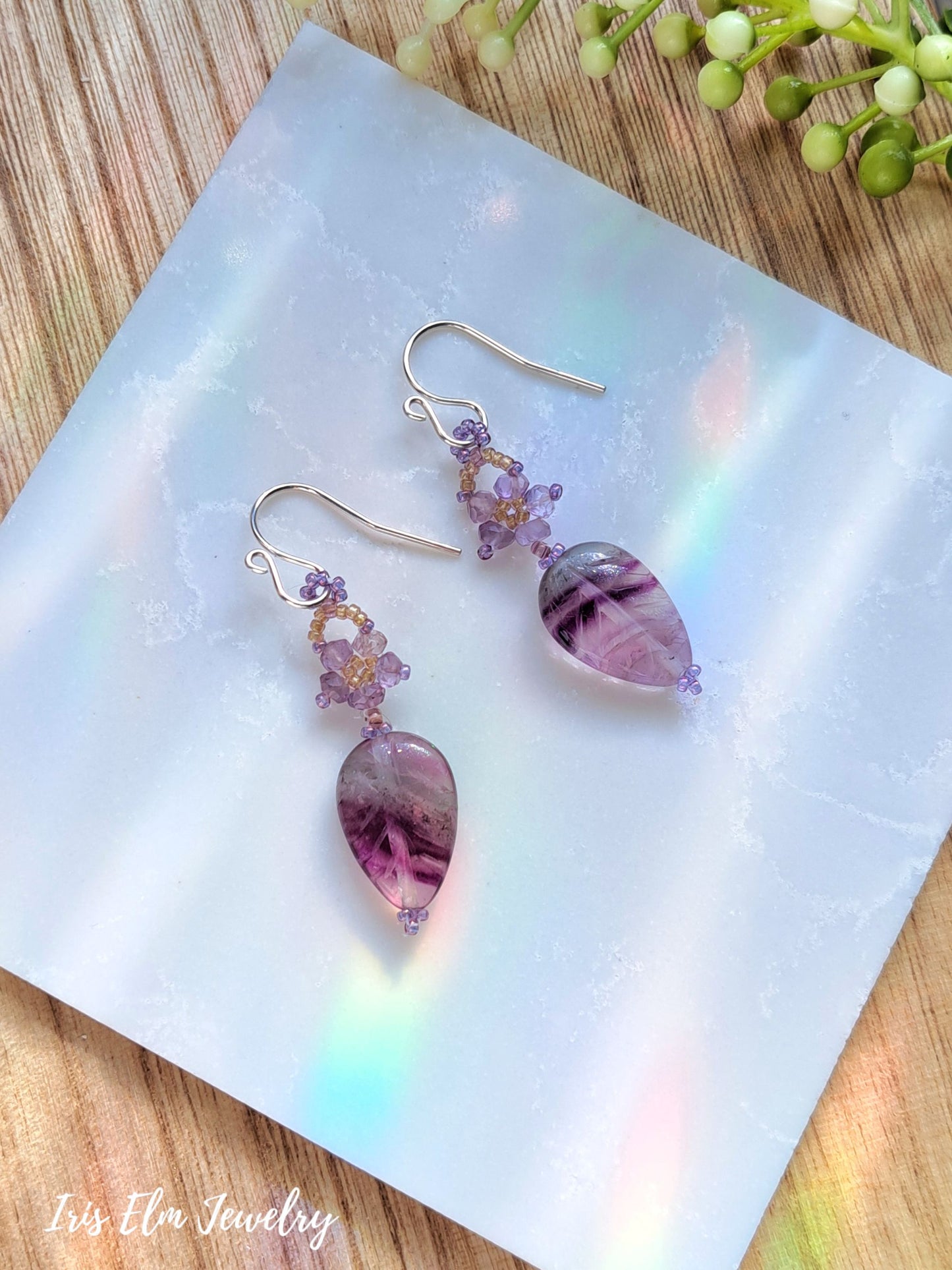 purple amethyst beaded flower and rainbow fluorite leaf gemstone dangle earrings on white marble surface