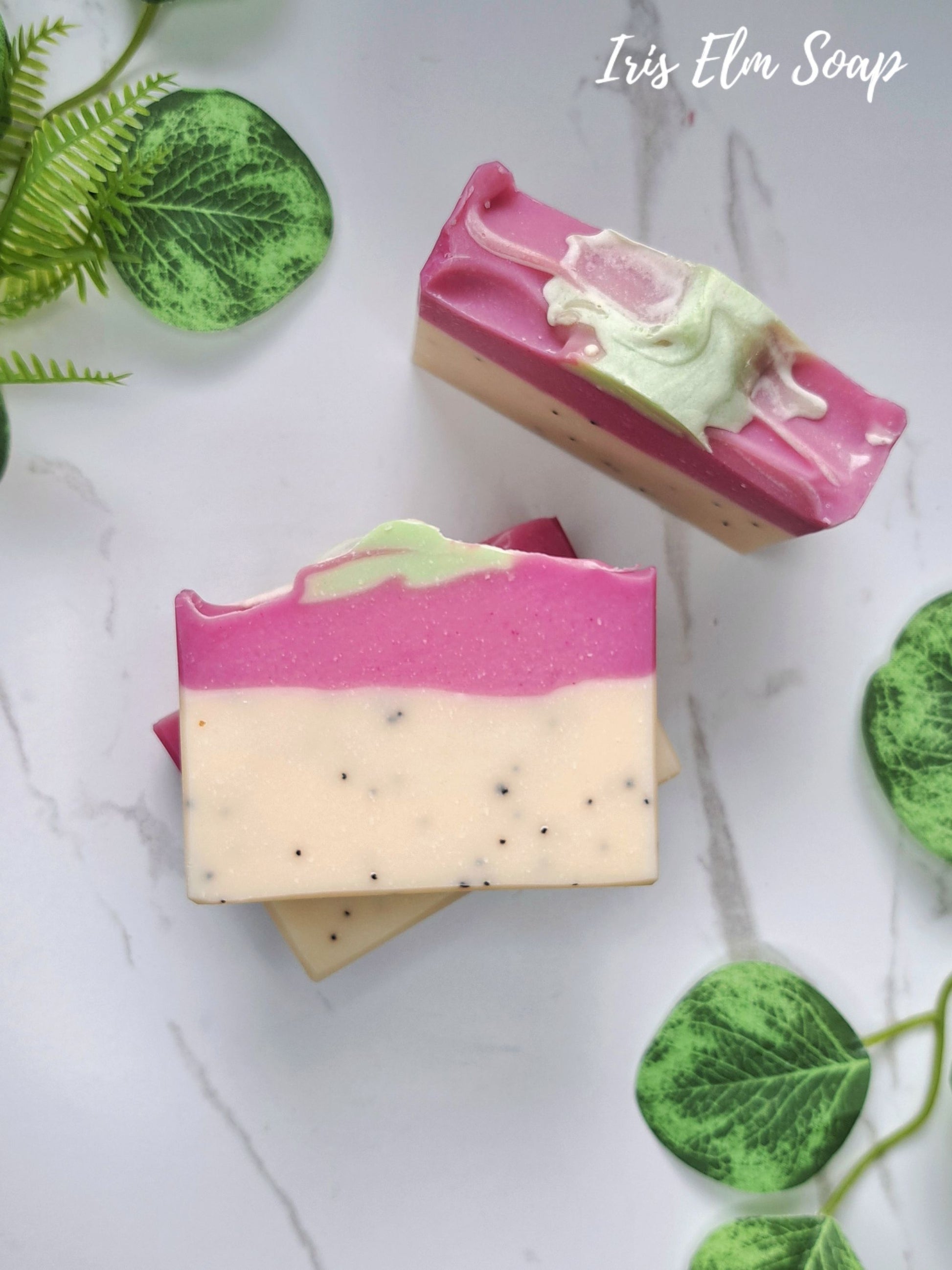 Two artisanal layered pink dragonfruit handmade soap bars on a white marble surface with decorative green leaves. The soaps feature a vibrant pink top layer, a pale cream middle layer with small black specks, and a light green swirl detail.