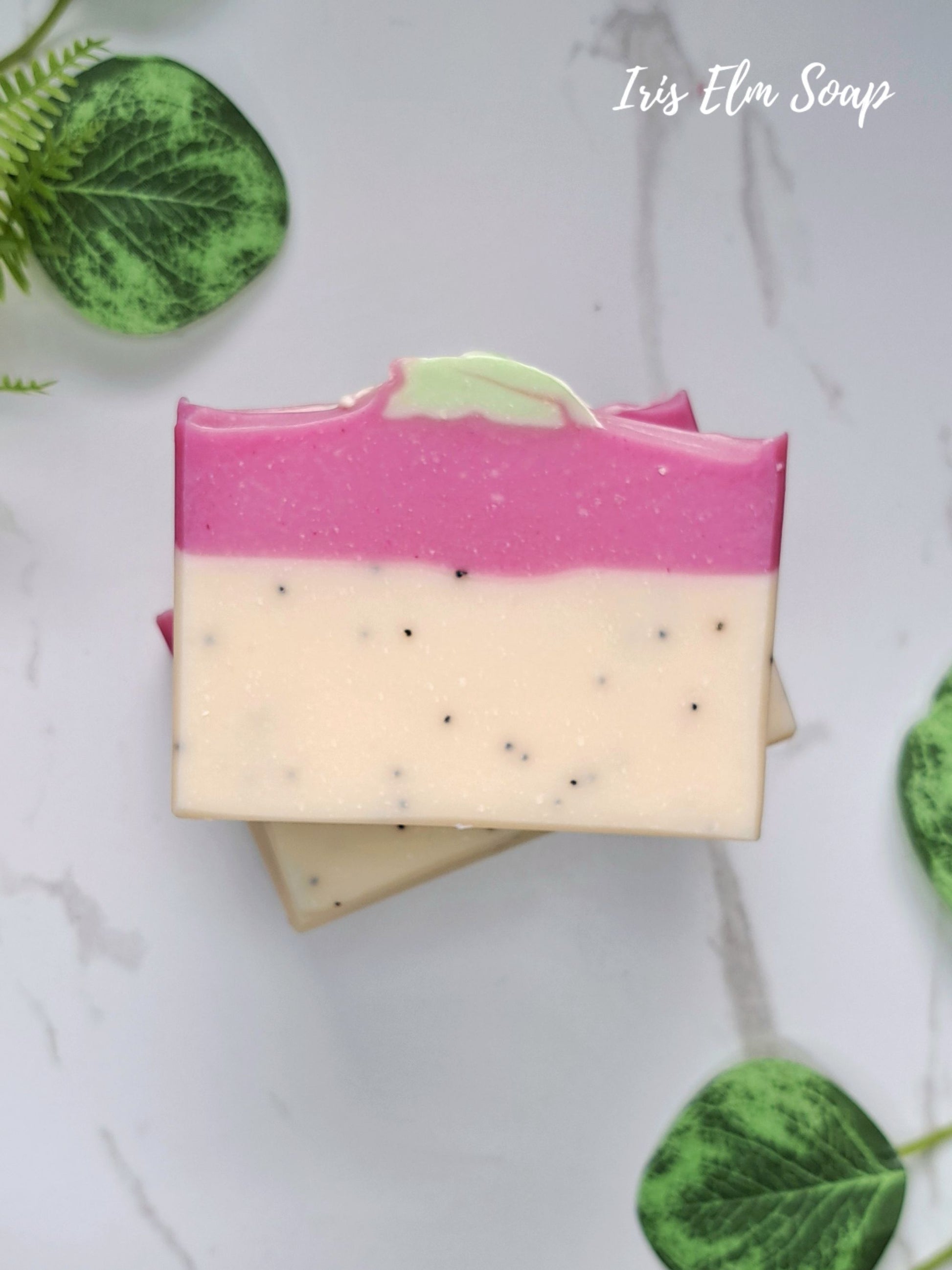 A handcrafted rectangular bar of pink dragonfruit soap with a two-tone design: a vibrant pink top layer and a creamy off-white bottom layer speckled with tiny black flecks. The soap is stacked on a white marble surface with decorative green leaves in the corners and the text 'Iris Elm Soap' in the top right corner.