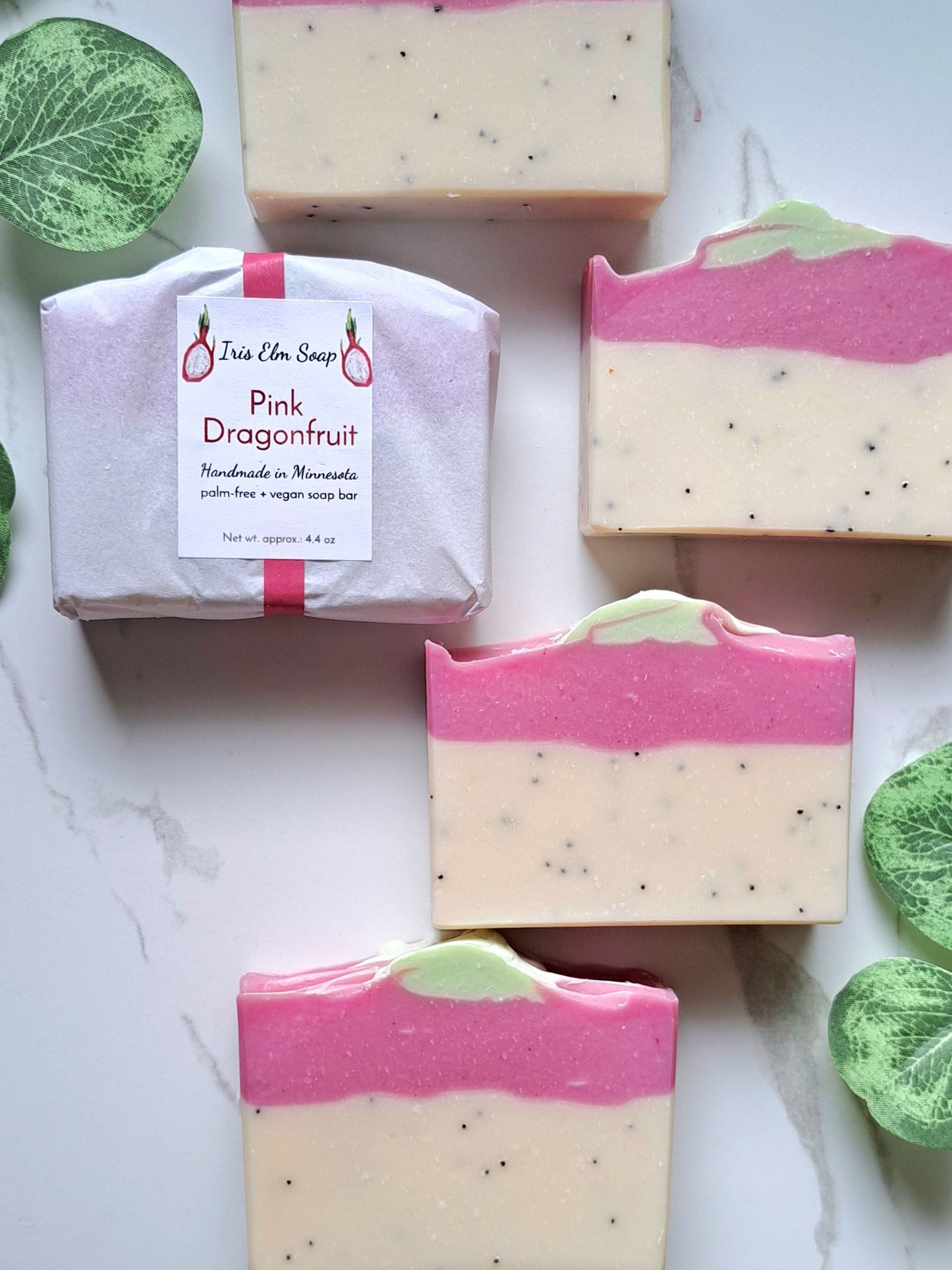 Five bars of artisan soap are arranged on a light marble surface with decorative green leaves. One soap bar is wrapped in pale pink paper with a white label reading 'Iris Elm Soap Pink Dragonfruit.