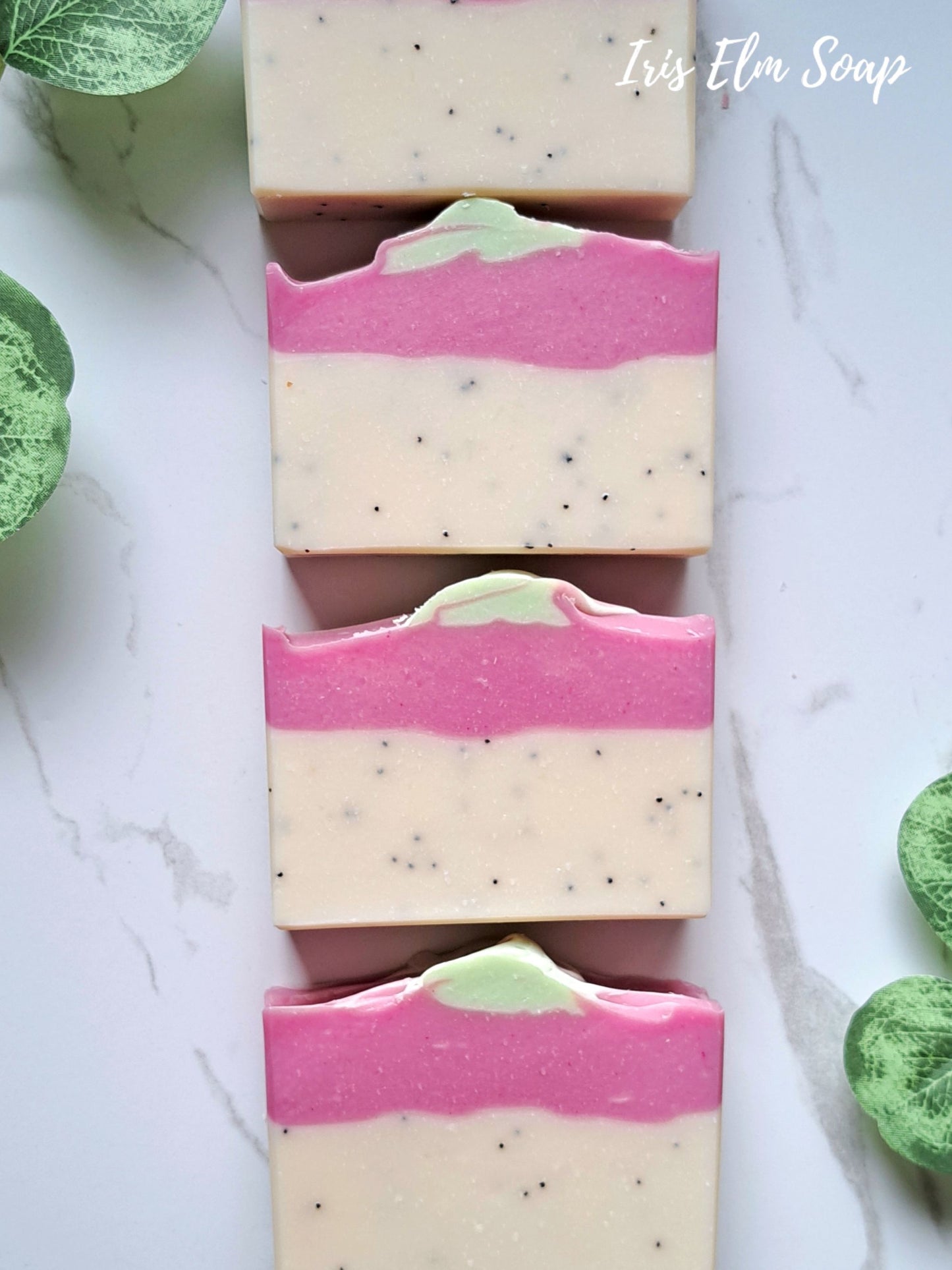 Four rectangular bars of pink dragonfruit tropical handmade soap with a cream-colored base, a vibrant pink middle band, and a small light green top swirl, arranged vertically on a white marble surface with green leaves at the corners. The text 'Iris Elm Soap' is visible in the top right corner.
