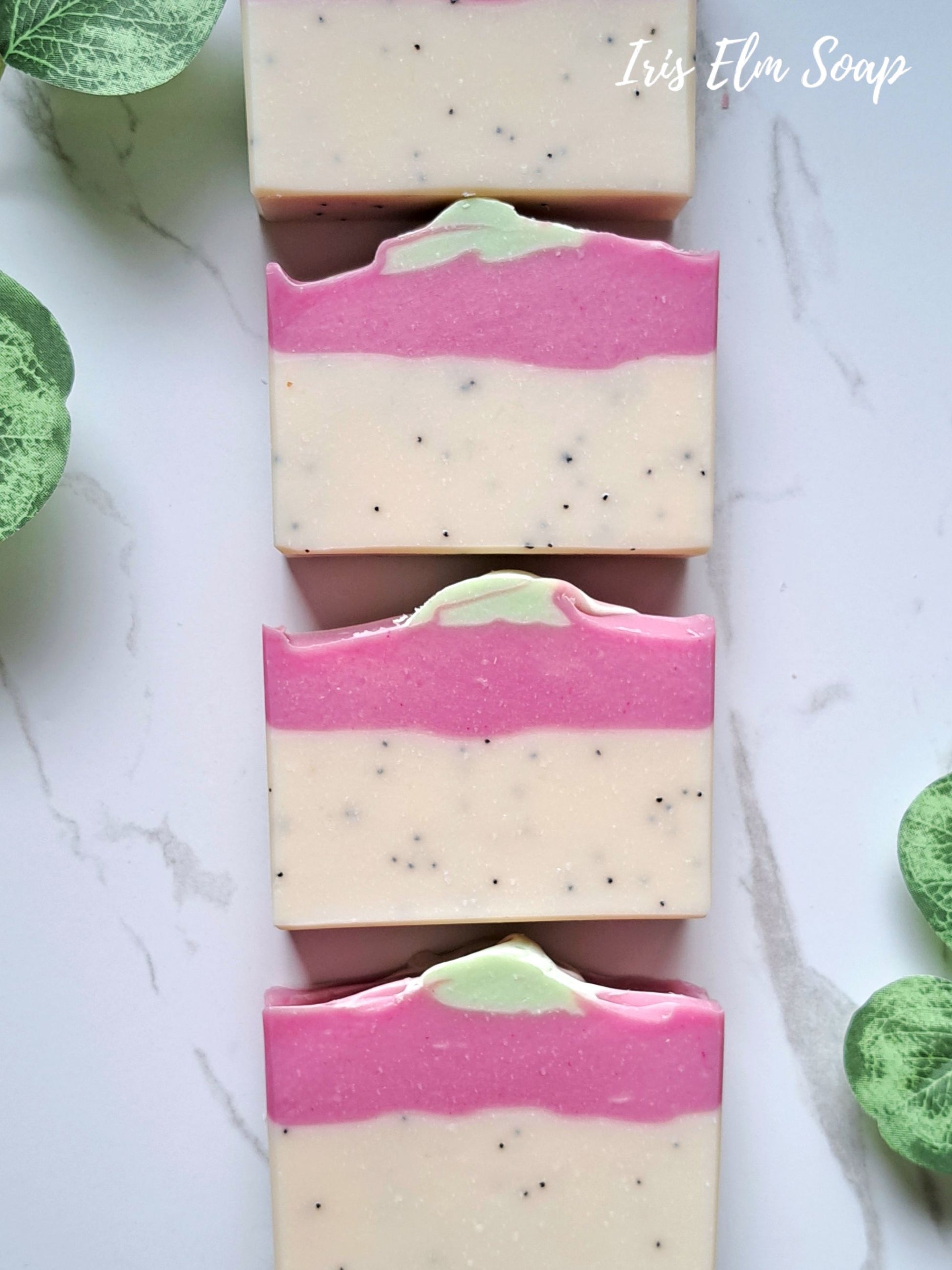 Four rectangular bars of pink dragonfruit tropical handmade soap with a cream-colored base, a vibrant pink middle band, and a small light green top swirl, arranged vertically on a white marble surface with green leaves at the corners. The text 'Iris Elm Soap' is visible in the top right corner.