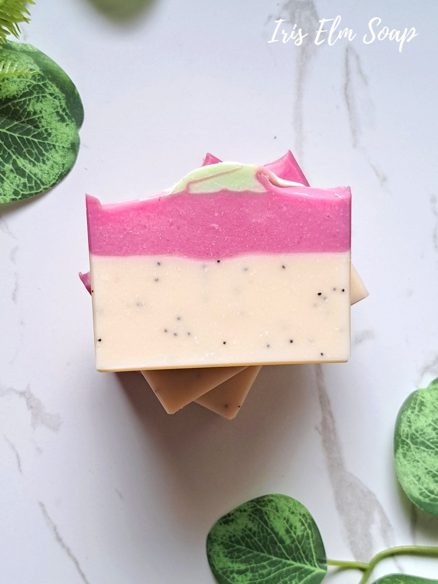A handcrafted rectangular bar of pink dragonfruit soap with a pink and cream marbled design, topped with a small green leaf, displayed on a white marble surface with green leaves around it. The text 'Iris Elm Soap' is visible in the top right corner.