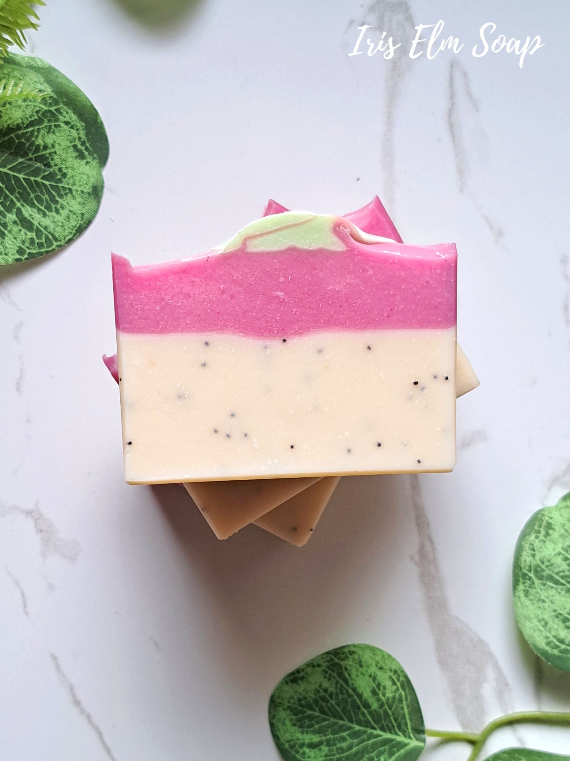 A handcrafted rectangular bar of pink dragonfruit soap with a pink and cream marbled design, topped with a small green leaf, displayed on a white marble surface with green leaves around it. The text 'Iris Elm Soap' is visible in the top right corner.