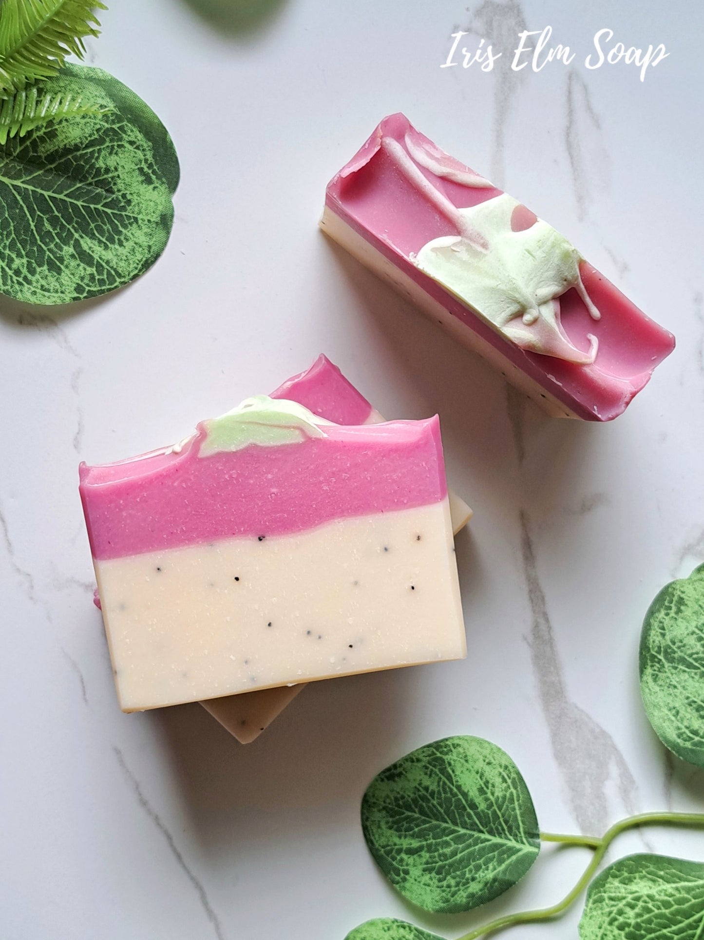 Two rectangular bars of layered handmade soap on a light marble surface with green leaf props. The soap has a bright pink top layer, a creamy off-white bottom layer with small dark specks, and a pale mint green swirled detail on top.