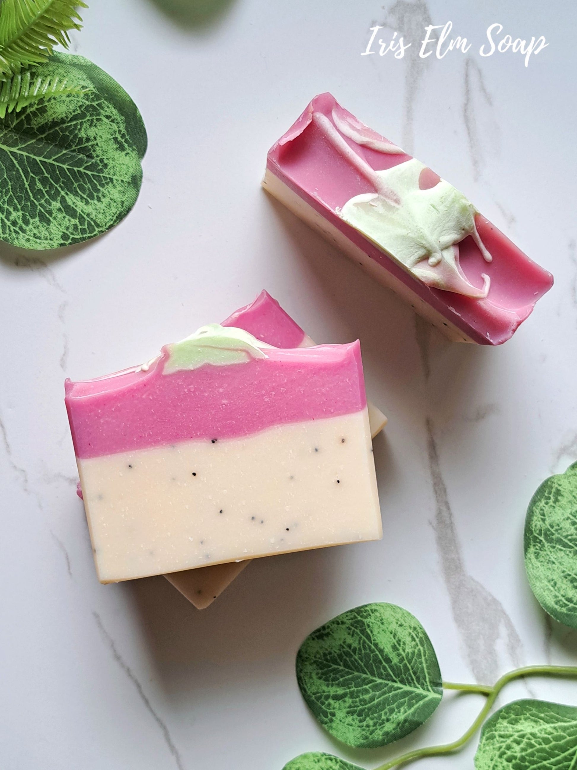 Two rectangular bars of layered handmade soap on a light marble surface with green leaf props. The soap has a bright pink top layer, a creamy off-white bottom layer with small dark specks, and a pale mint green swirled detail on top.