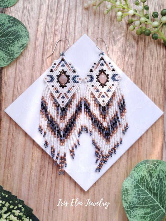Beaded diamond seed bead fringe celestial peach moonstone earrings on a white card with green leaves around, Iris Elm Jewelry branding visible.