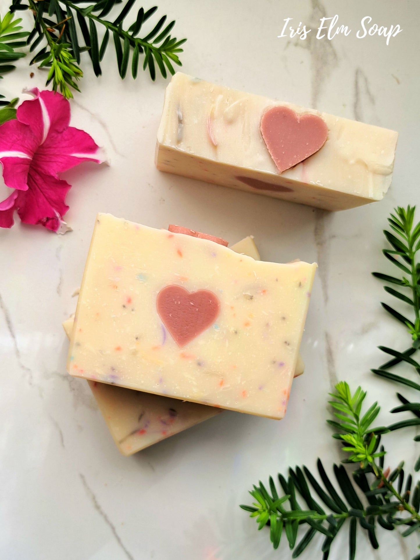 Three bars of persephone's kiss handmade spring goddess soap with heart designs on a white surface with greenery and flowers.