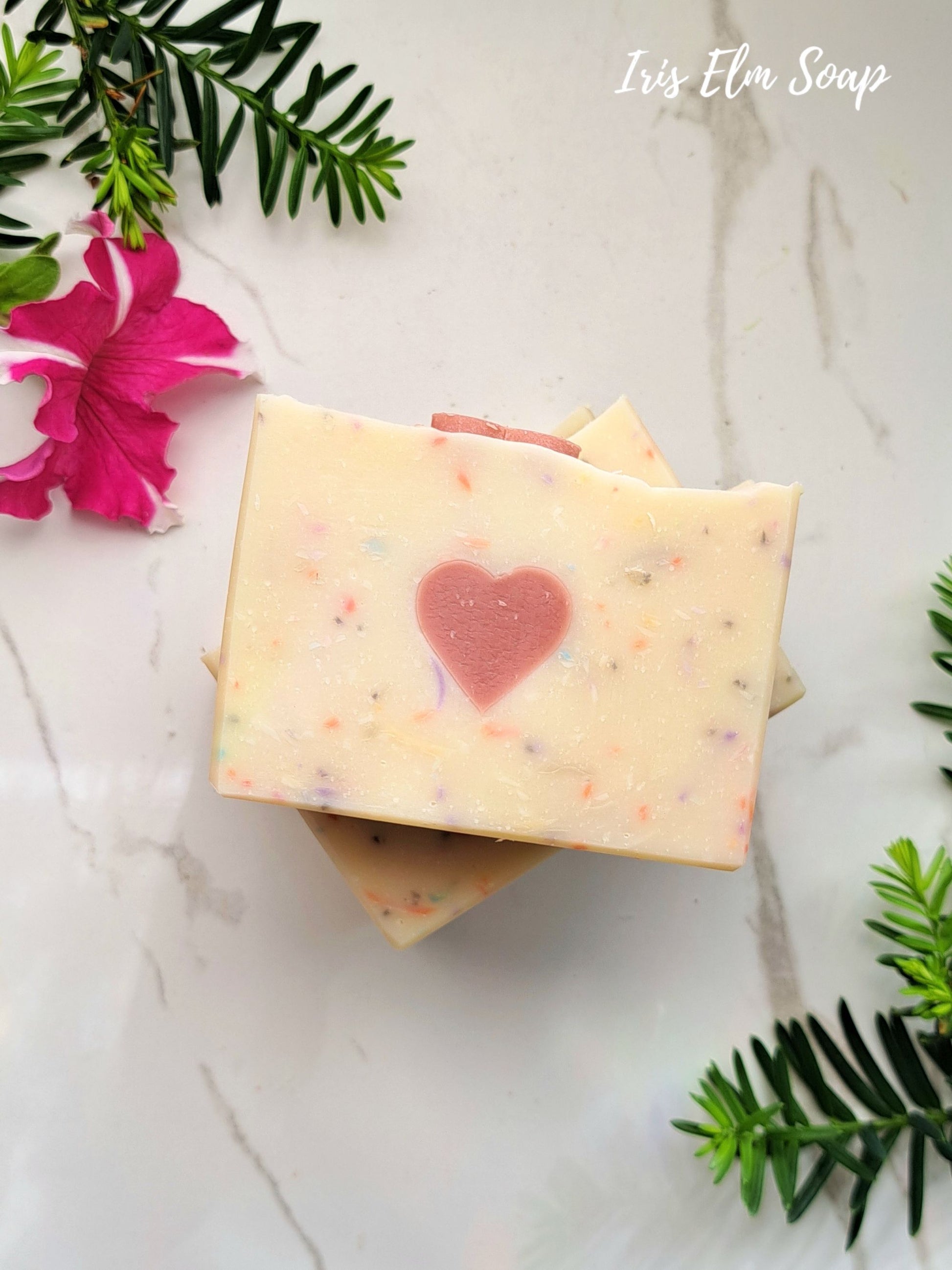 Two persephone's kiss handmade spring goddess soap bars with a heart design on a marble surface with greenery and flowers.