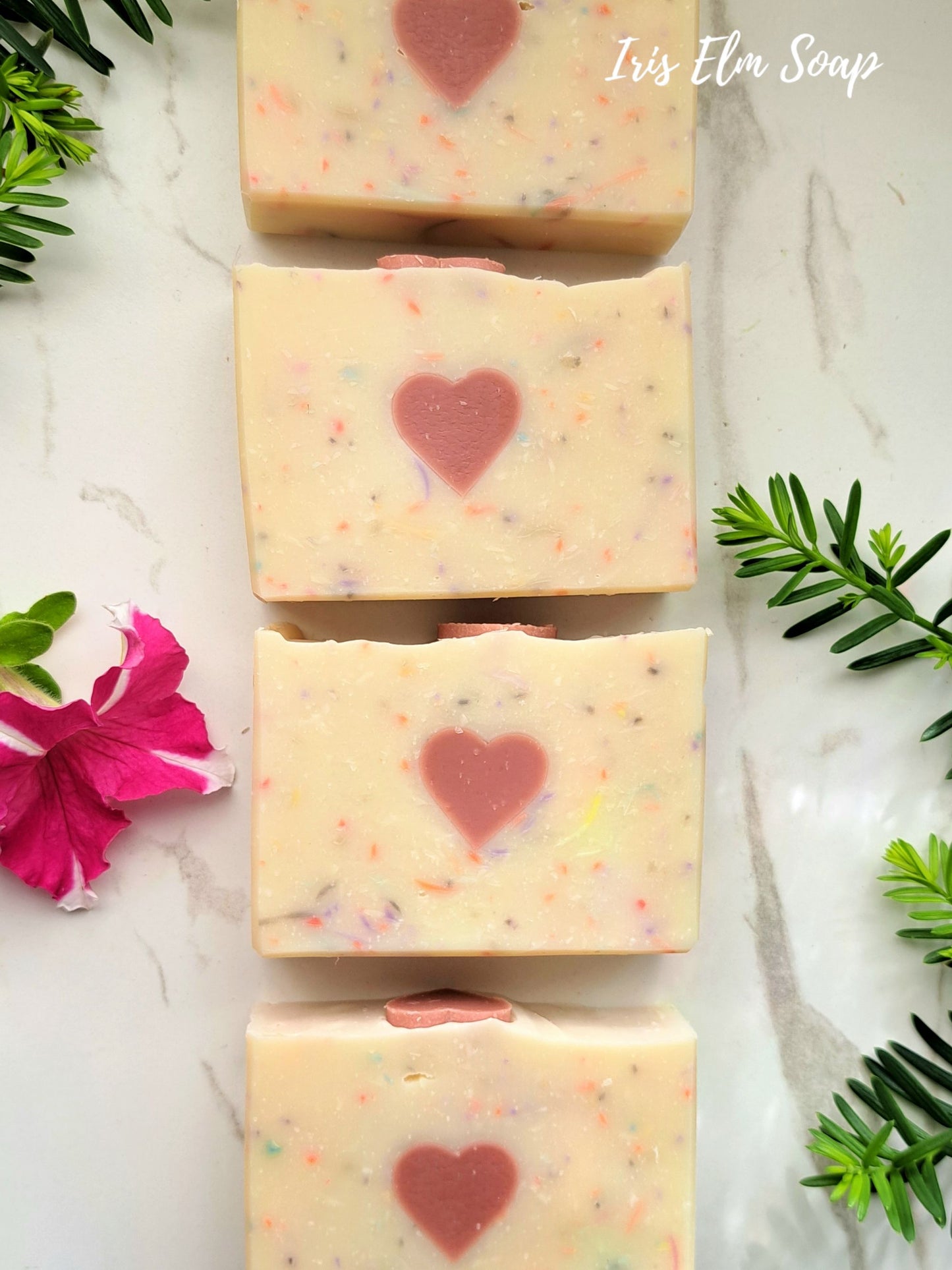 Four persephone's kiss handmade spring goddess soap bars with heart designs on a marble surface with greenery and flowers.