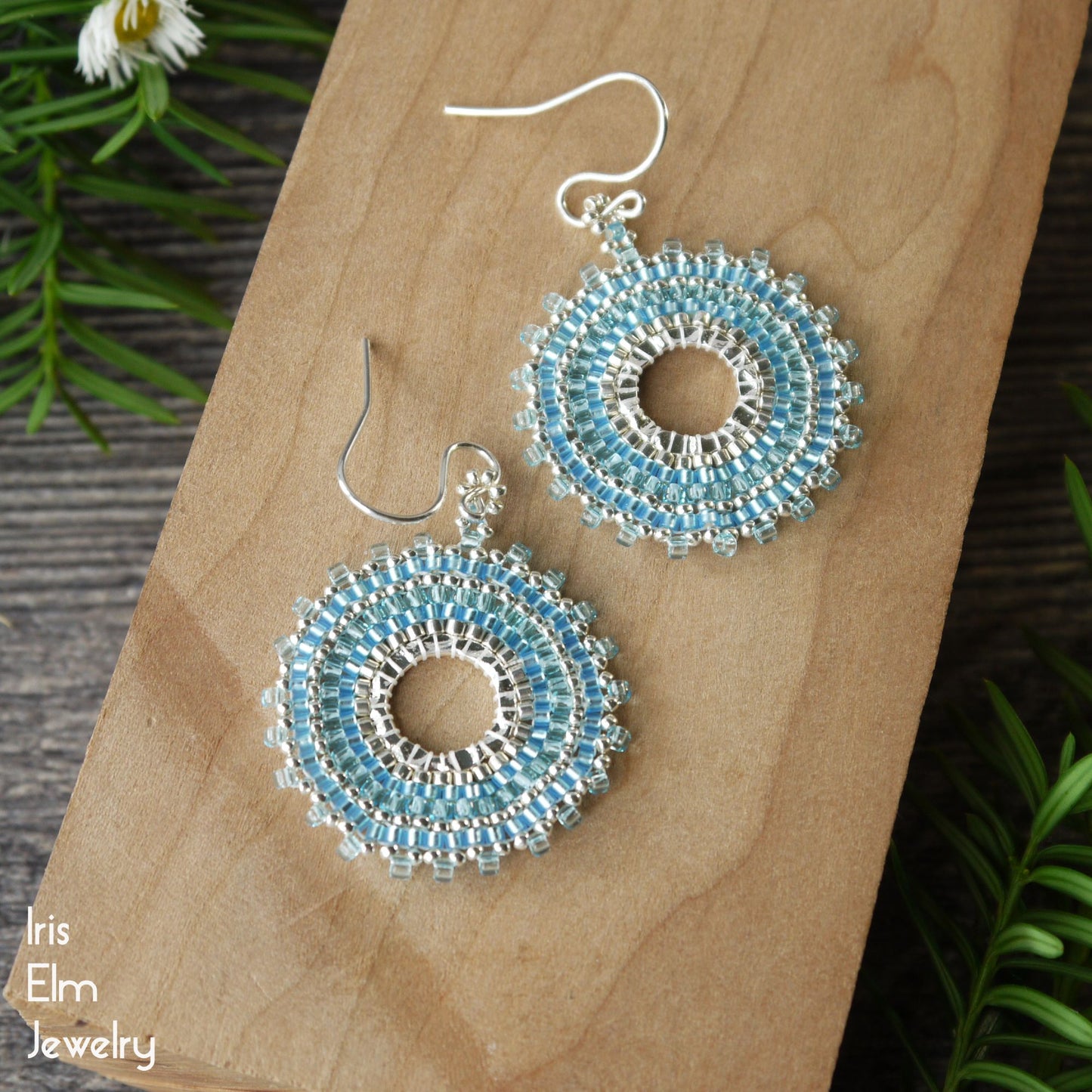 Circular blue and silver handmade beaded earrings with sterling silver ear wires on a wooden surface with 'Iris Elm Jewelry' branding.