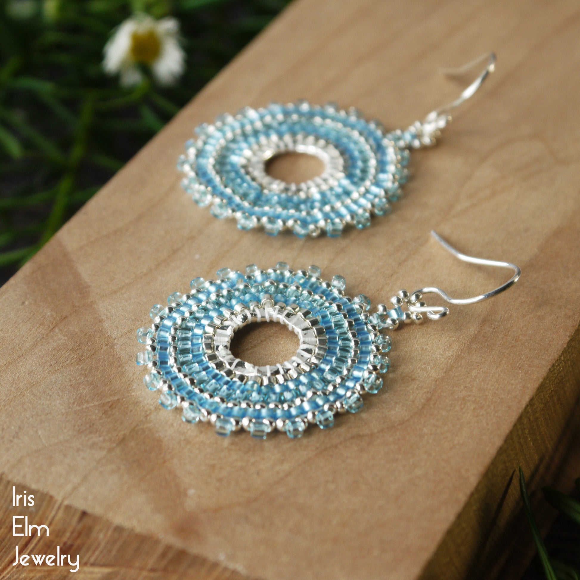 Pair of light blue seed bead beaded hoop earrings on a wooden surface with 'Iris Elm Jewelry' branding.