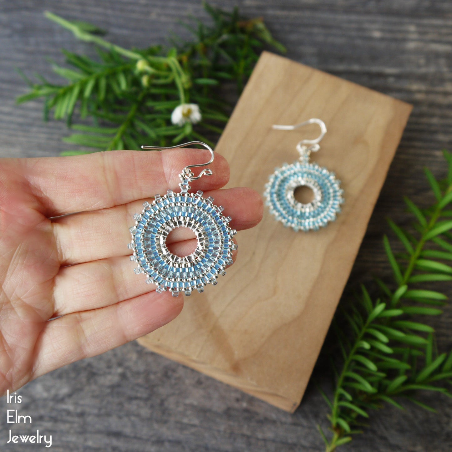 Hand holding a light blue glass seed bead pair of beaded woven hoop earrings with a wooden card and greenery in the background