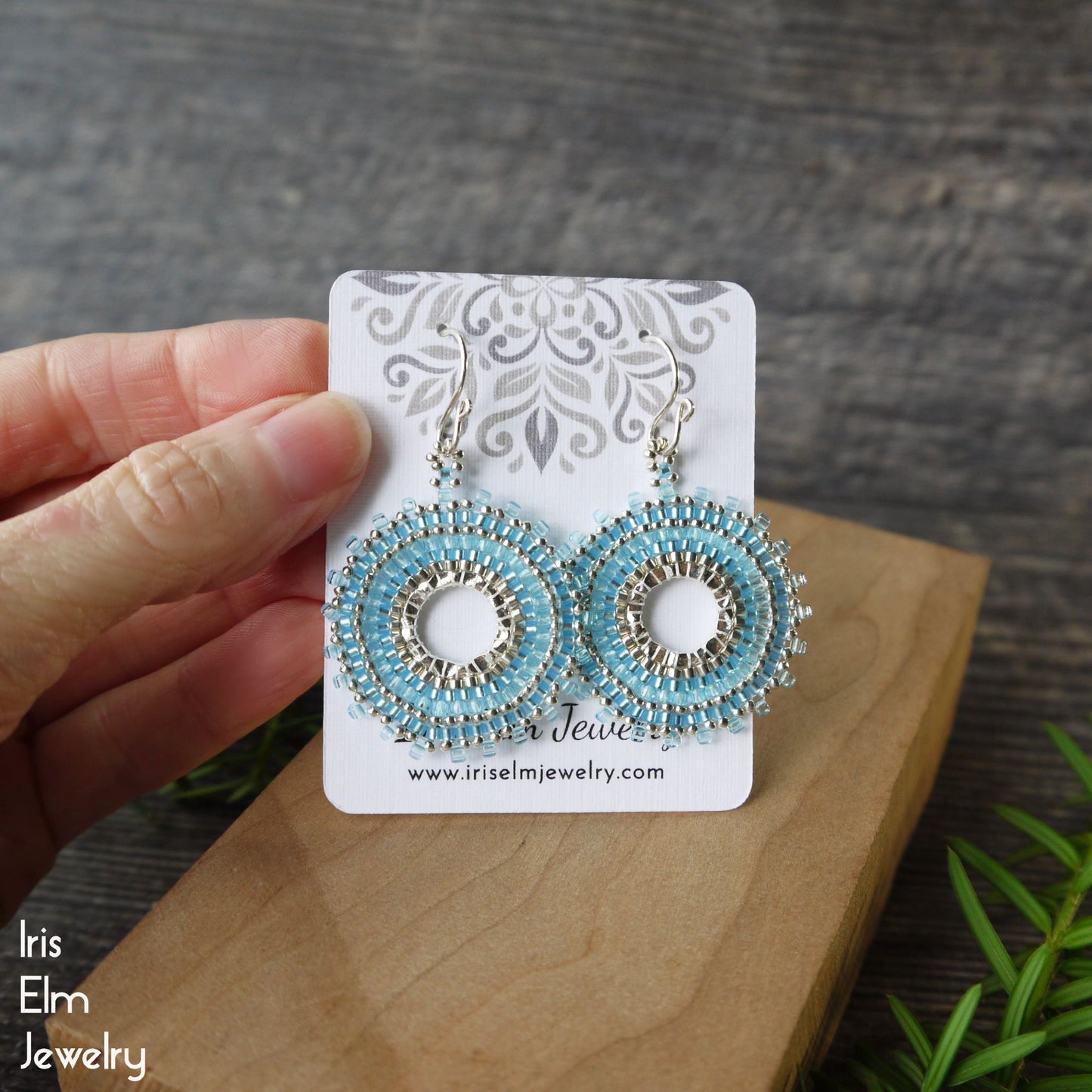 Blue beaded circular woven hoop seed bead earrings on a card held by a hand with a wooden surface and plant in the background