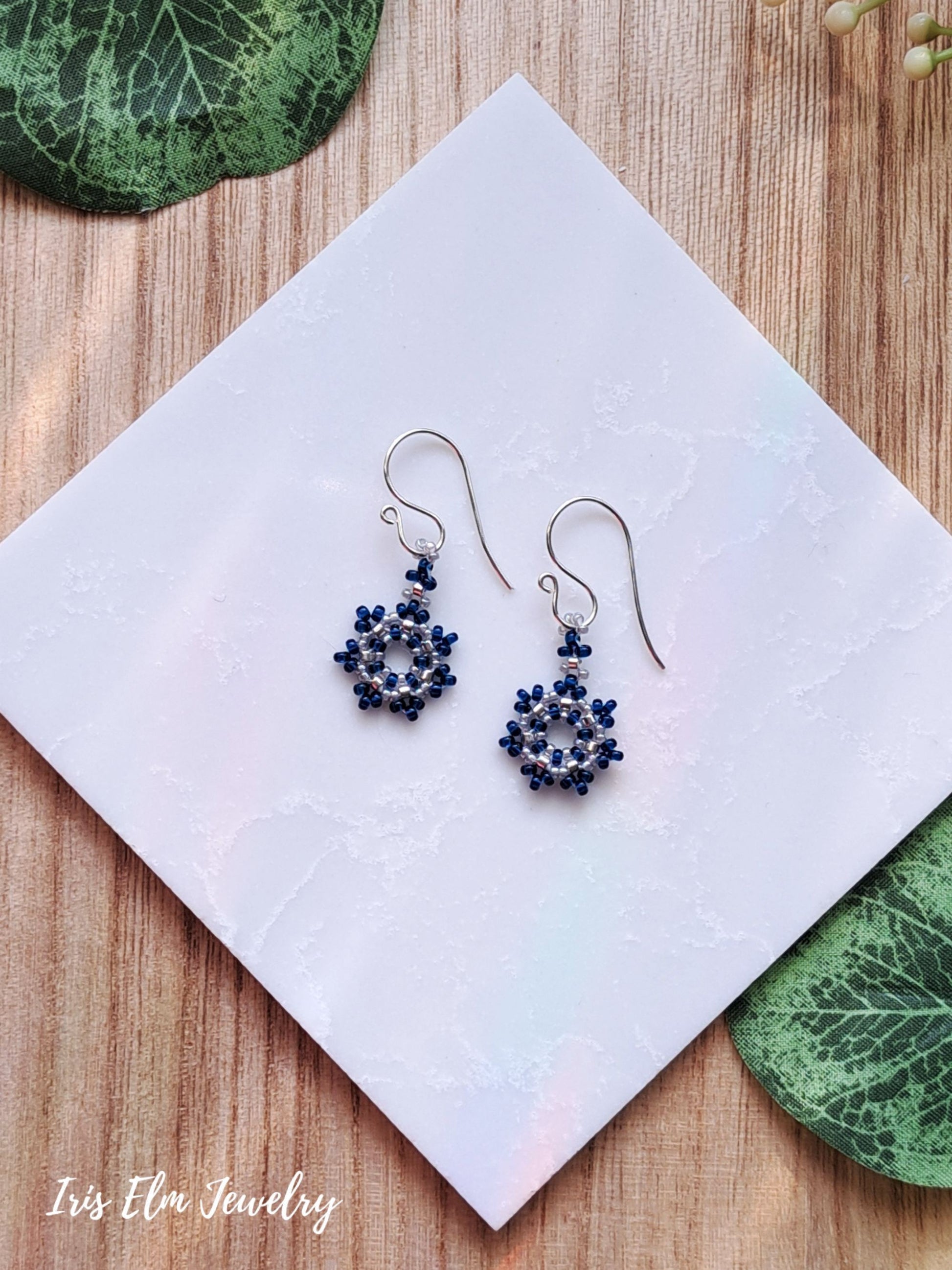 Pair of cobalt blue beaded seed bead snowflake earrings on a white tile with green leaves in the background