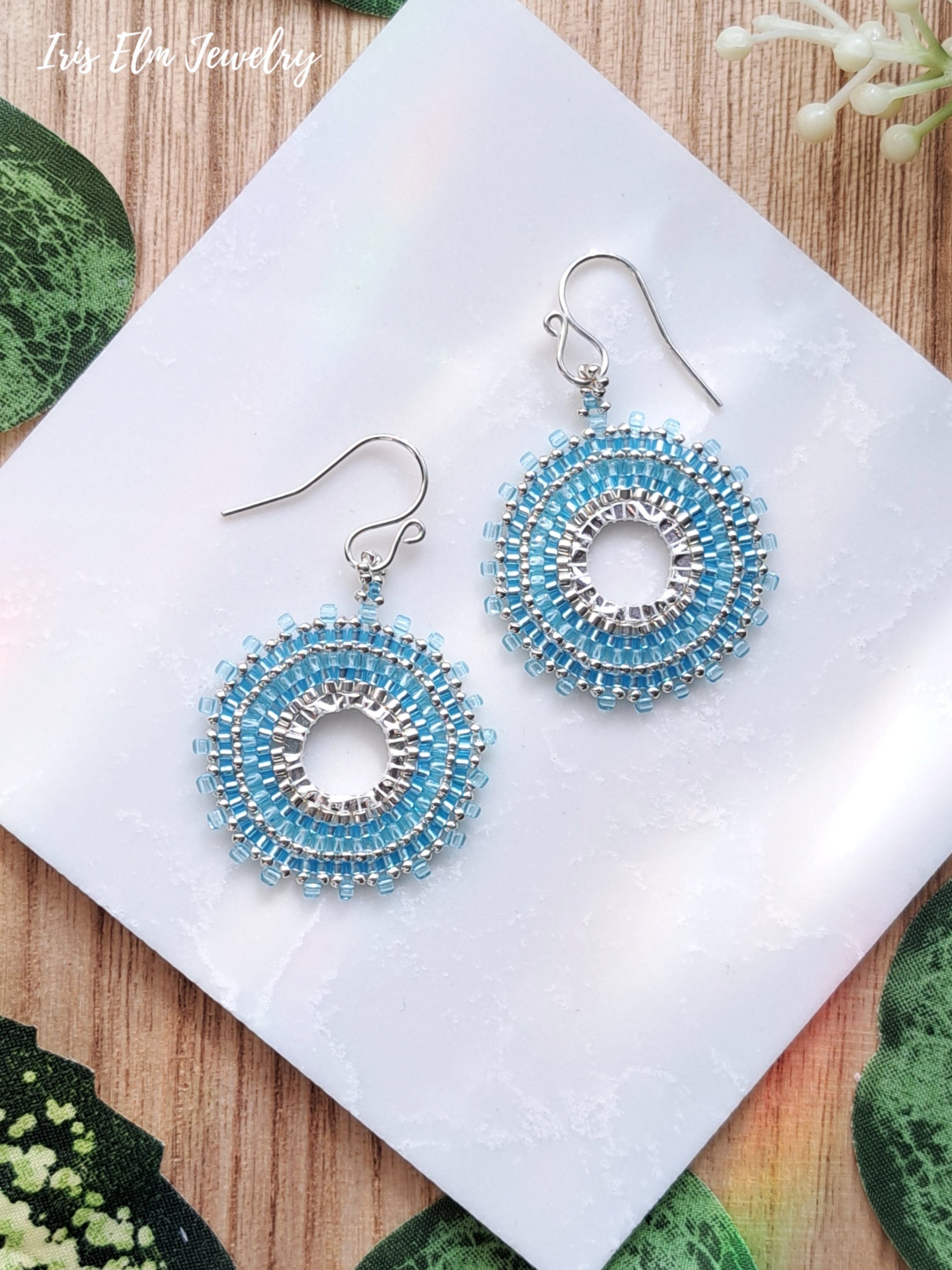 Pair of beaded light blue seed bead woven hoop earrings on a white card with a wooden background