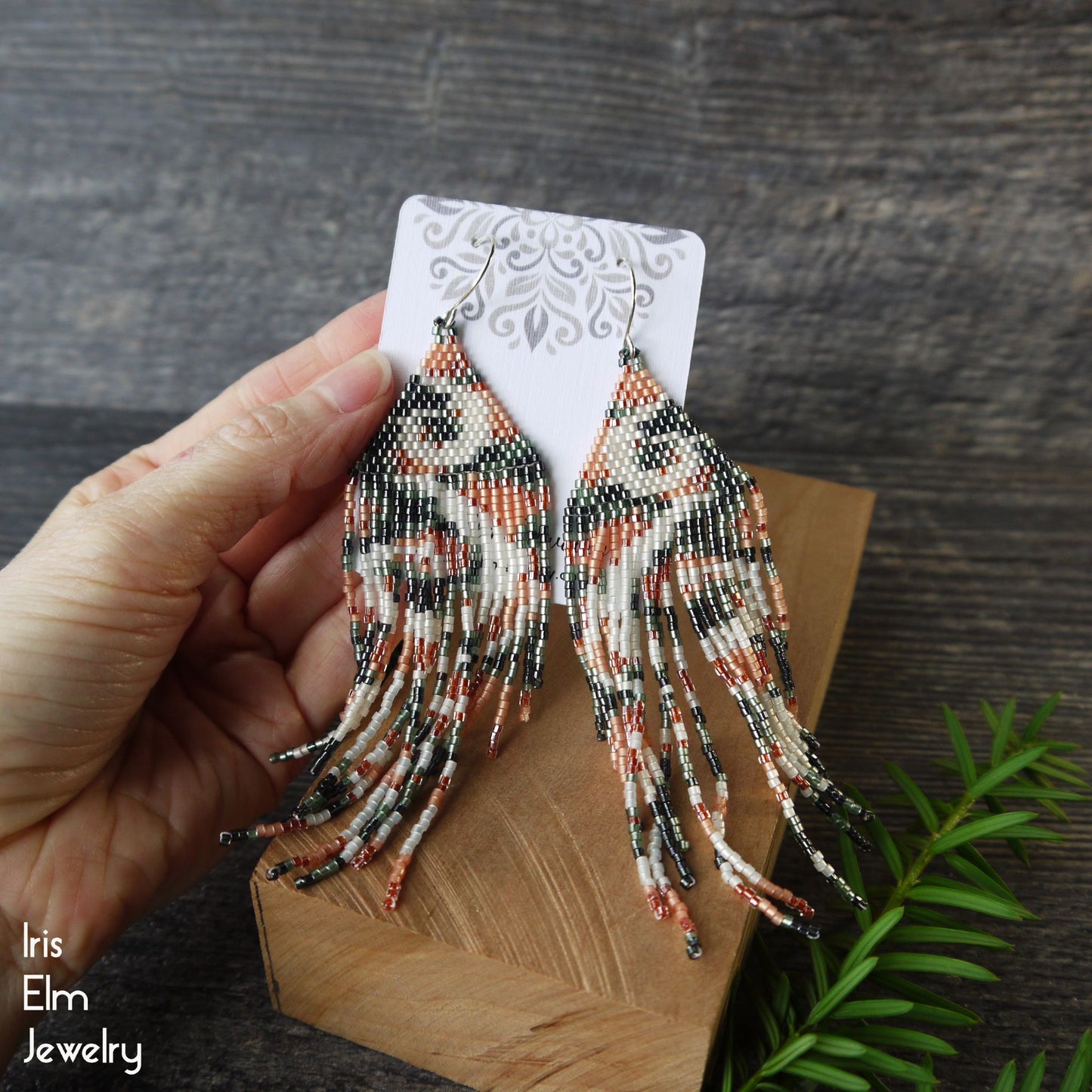 Beaded seed bead fringe swirl patterned earrings held by a hand on a wooden stand with a dark background