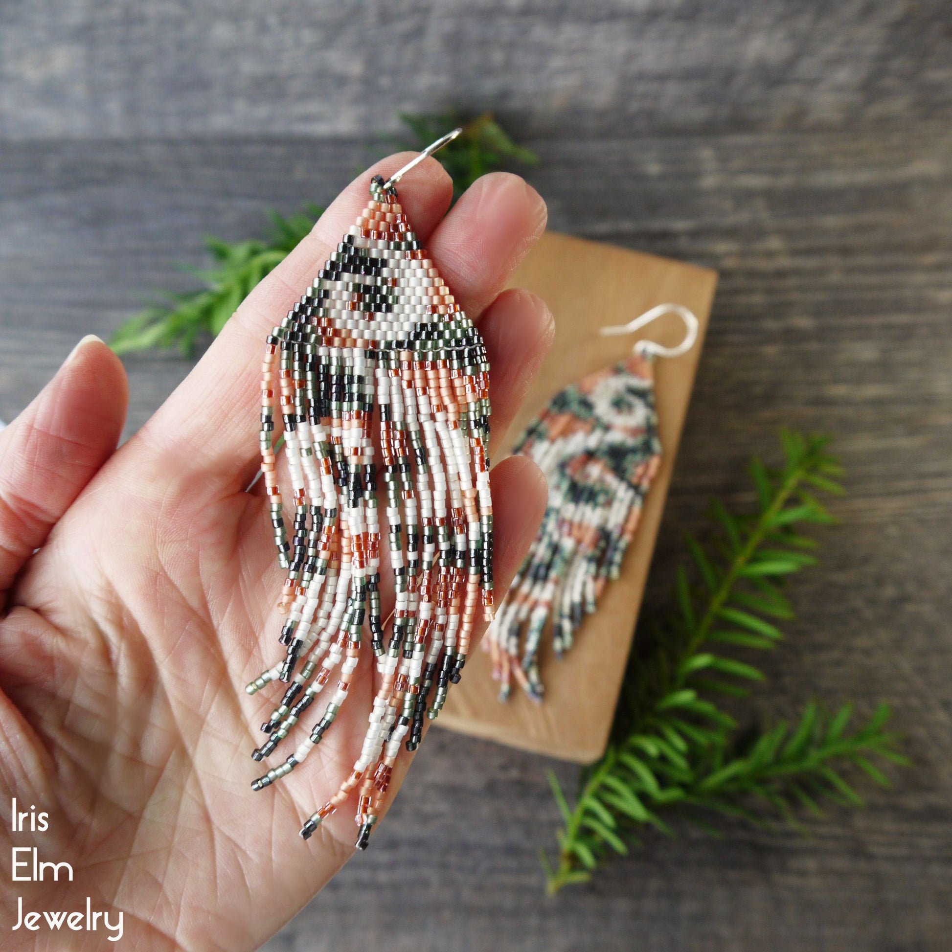 Hand holding long beaded peach green and white seed bead fringe swirl earrings with a wooden box and greenery in the background, featuring 'Iris Elm Jewelry'.