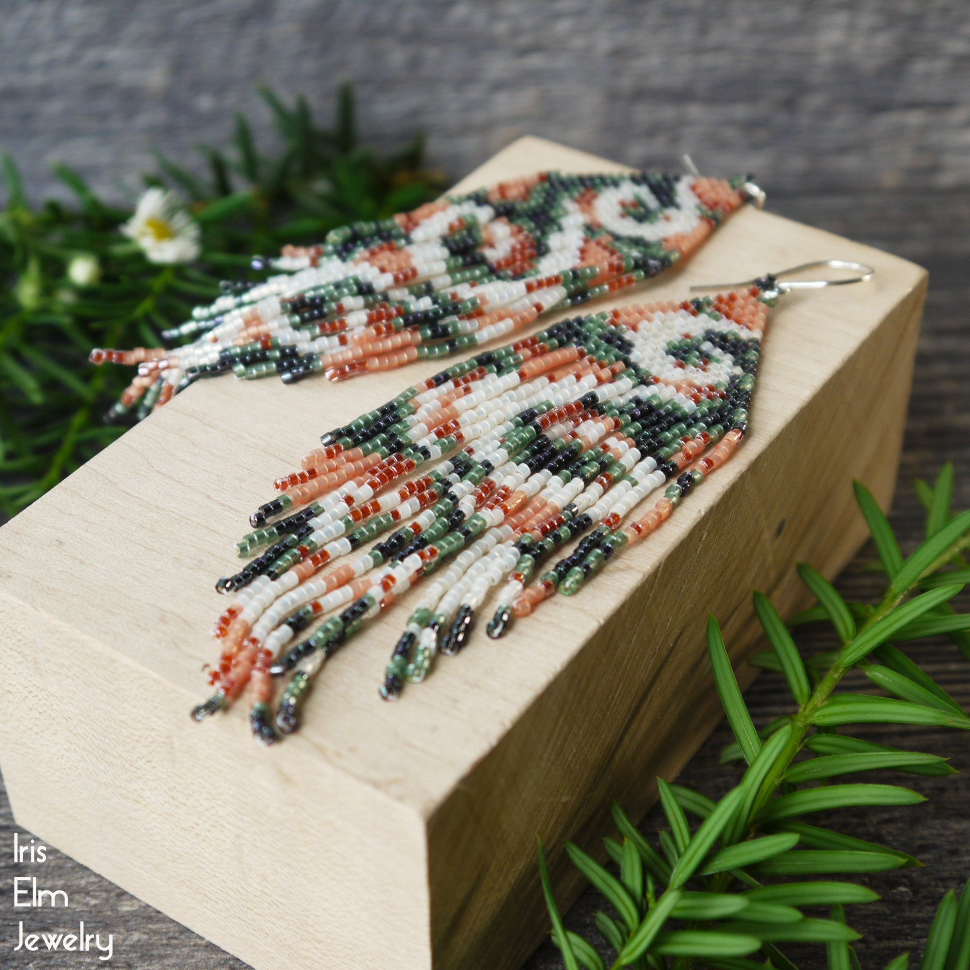 Beaded green and peach seed bead fringe earrings on a wooden block with greenery in the background