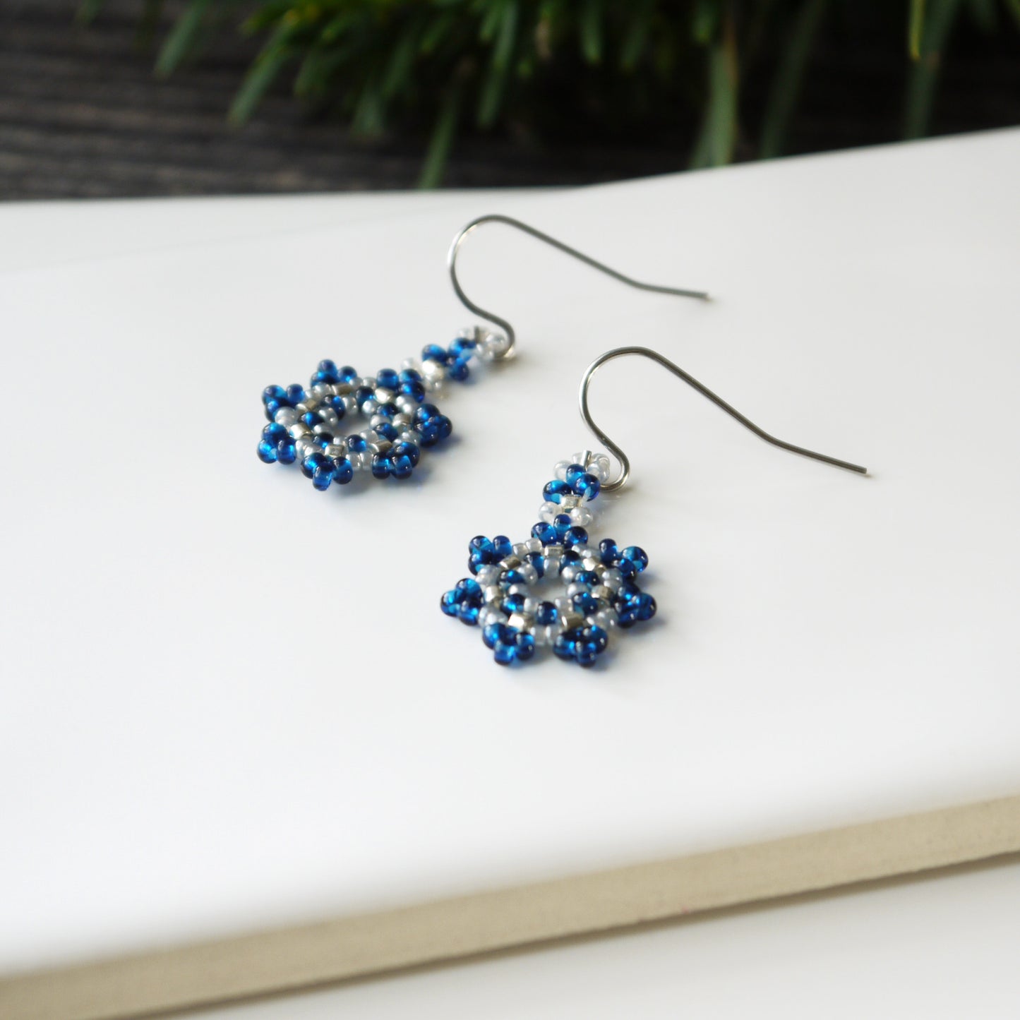 Handmade blue snowflake beadwork earrings on a white surface with a blurred natural background