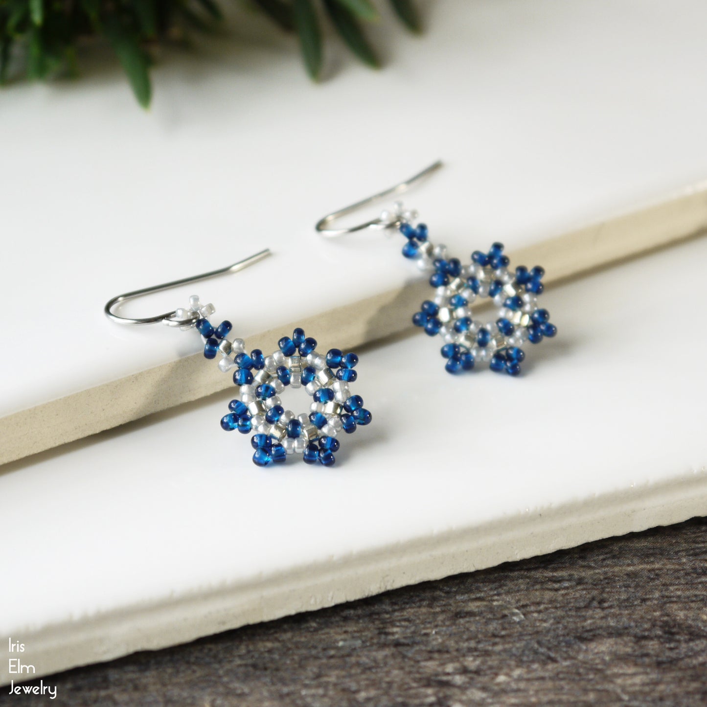 Pair of cobalt blue and silver beaded winter snowflake earrings on a white surface with greenery in the background.
