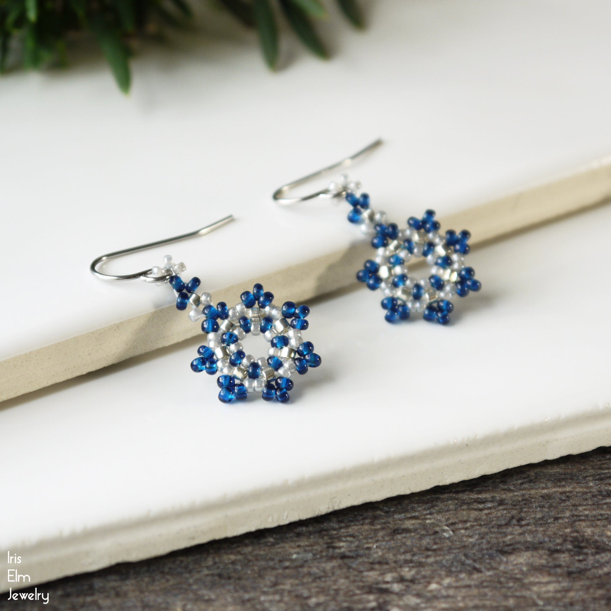 Pair of cobalt blue and silver beaded winter snowflake earrings on a white surface with greenery in the background.