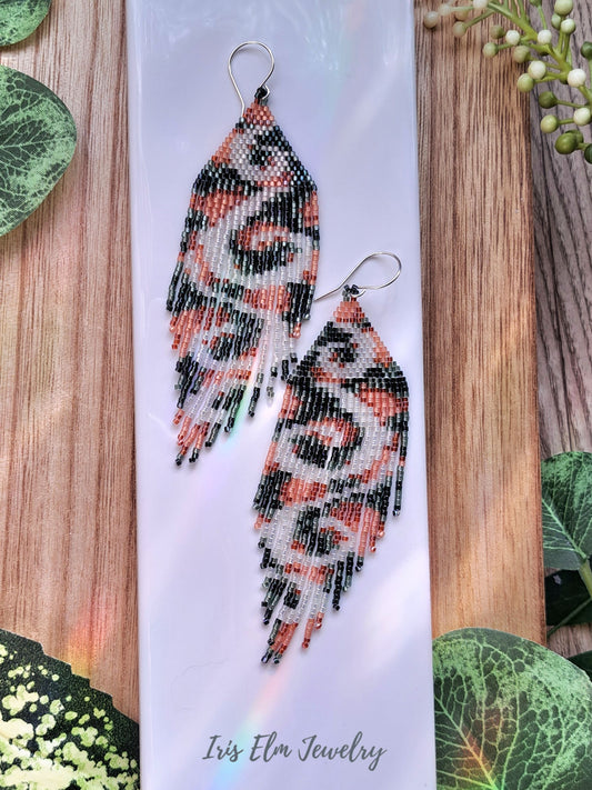 Beaded peach green white swirl seed bead fringe earrings on a white card with 'Iris Elm Jewelry' branding, surrounded by green leaves on a wooden surface.